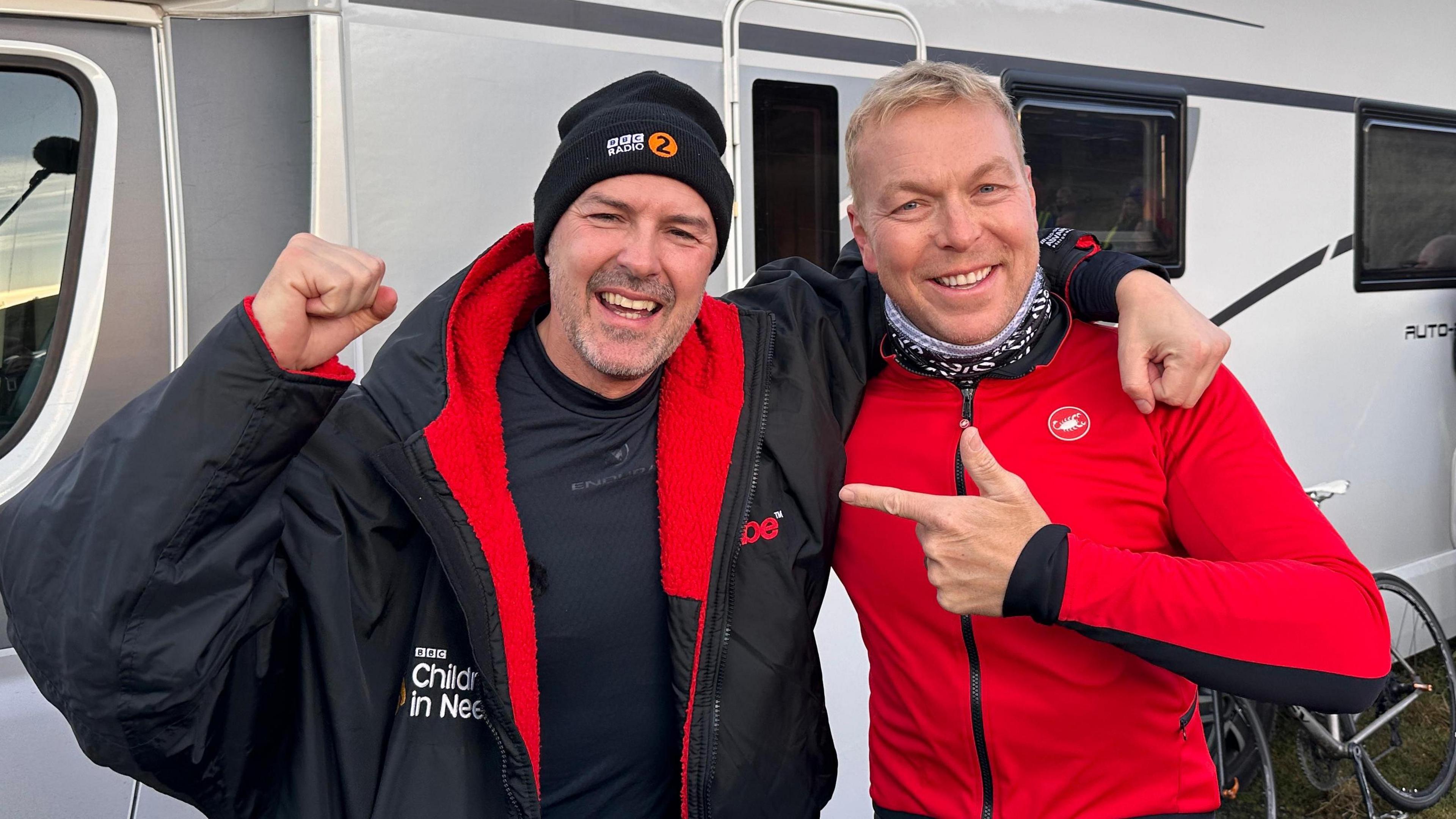 Paddy McGuinness said Sir Chris Hoy outside a campervan. Paddy wears black hat and pumps his first in the air and chris is in a red cycling top pointing at paddy.