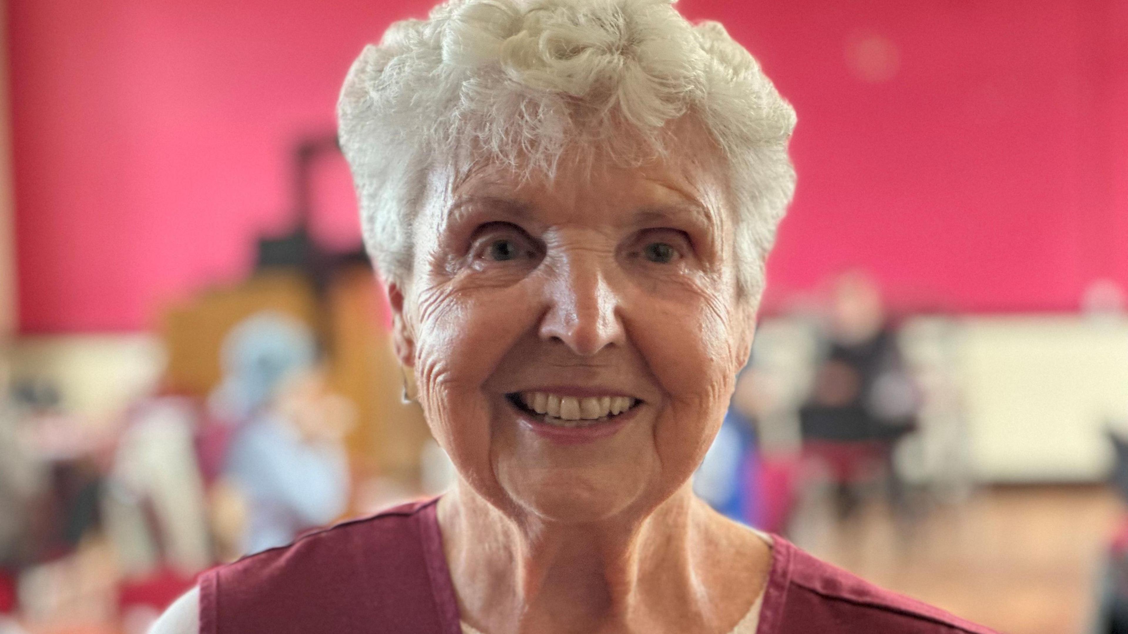 Carol Fox, who has short curly white hair, smiling. She is wearing a maroon pinafore over a white top.