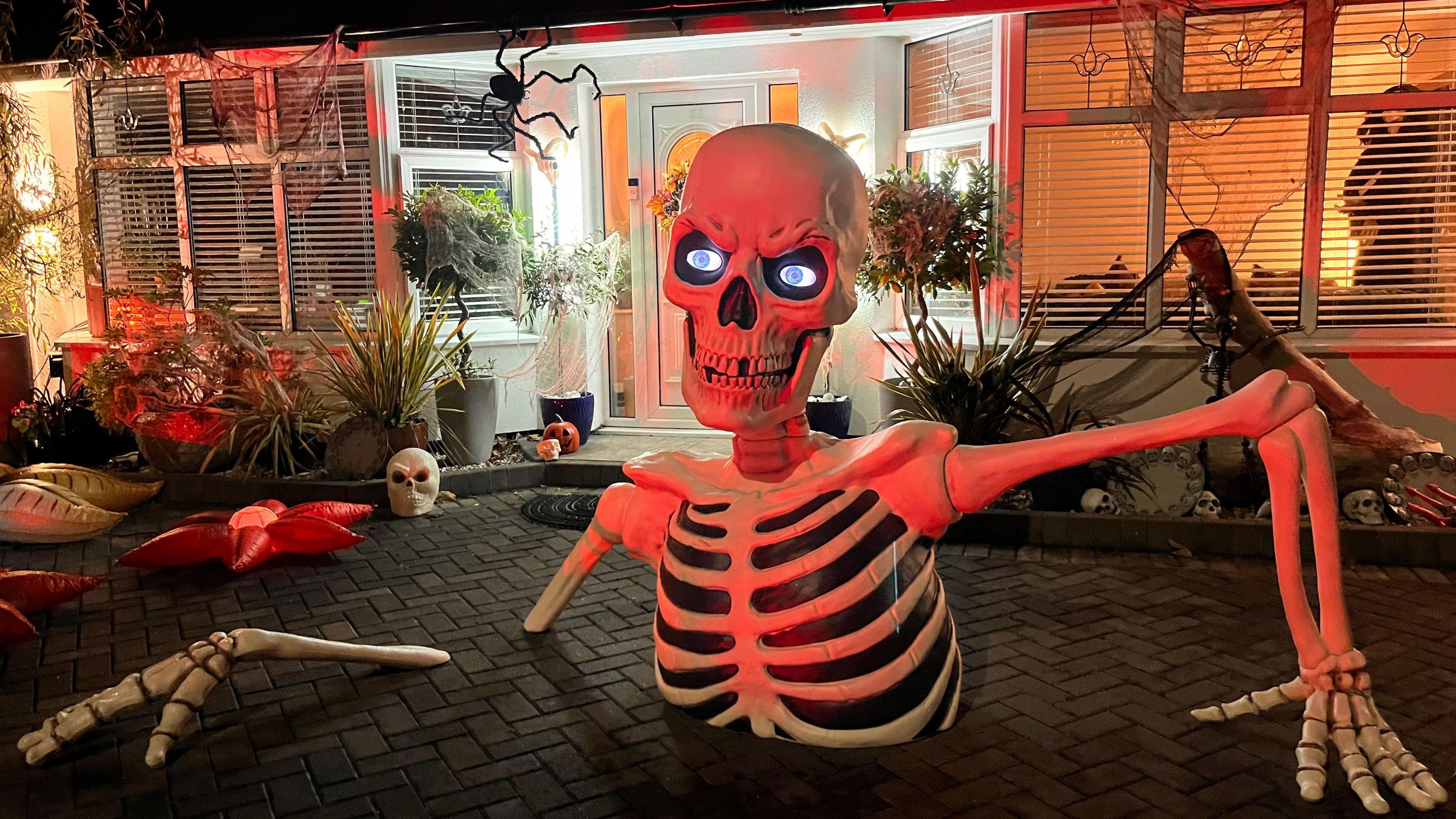A large skeleton appears to be coming up through the ground at the front of a bungalow, with other Halloween decorations such as spiders and spider webs all around.