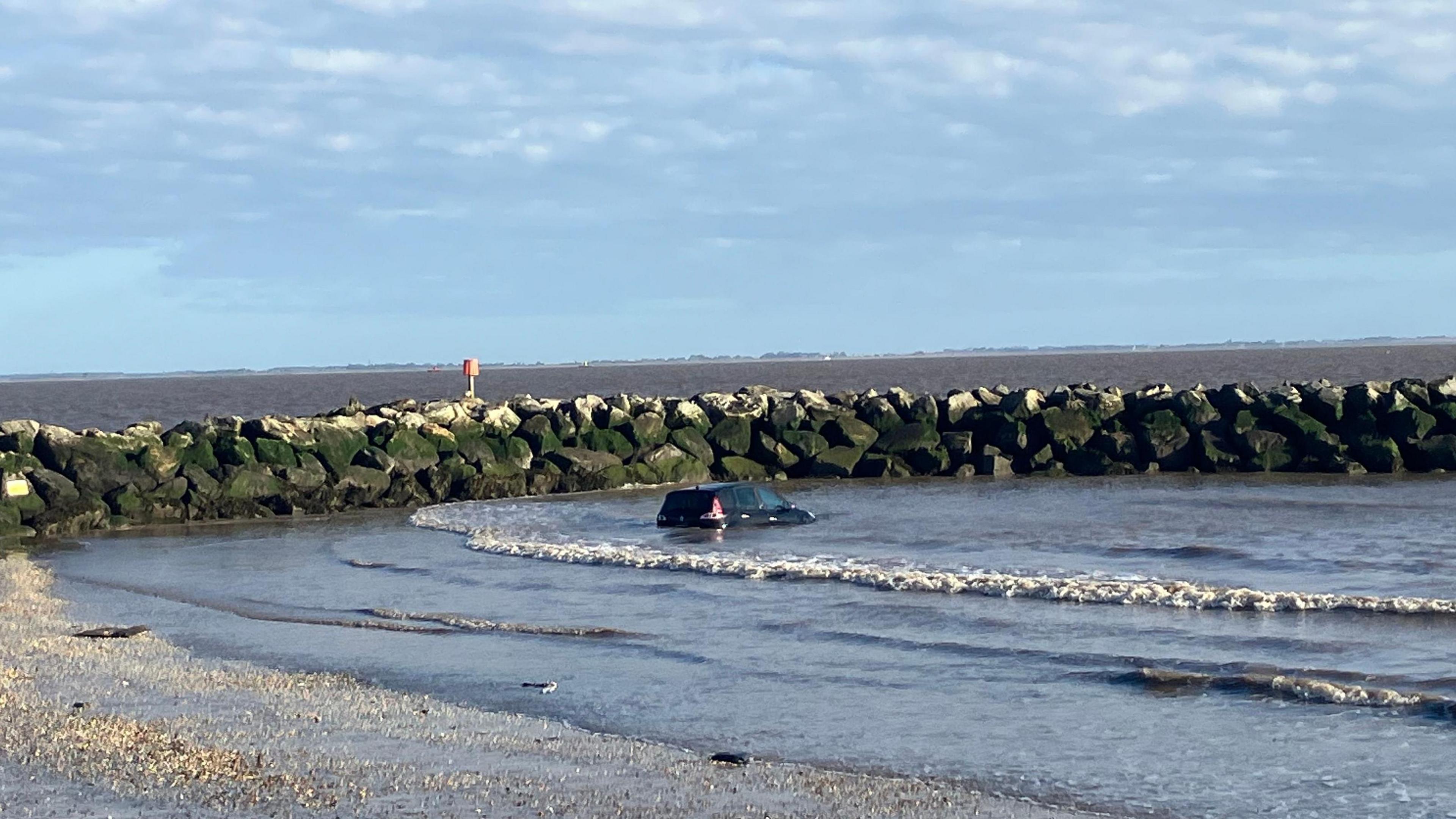 A black car can be seen submerged by waves next to a sea wall of rocks. The beach can be seen in the foreground. 