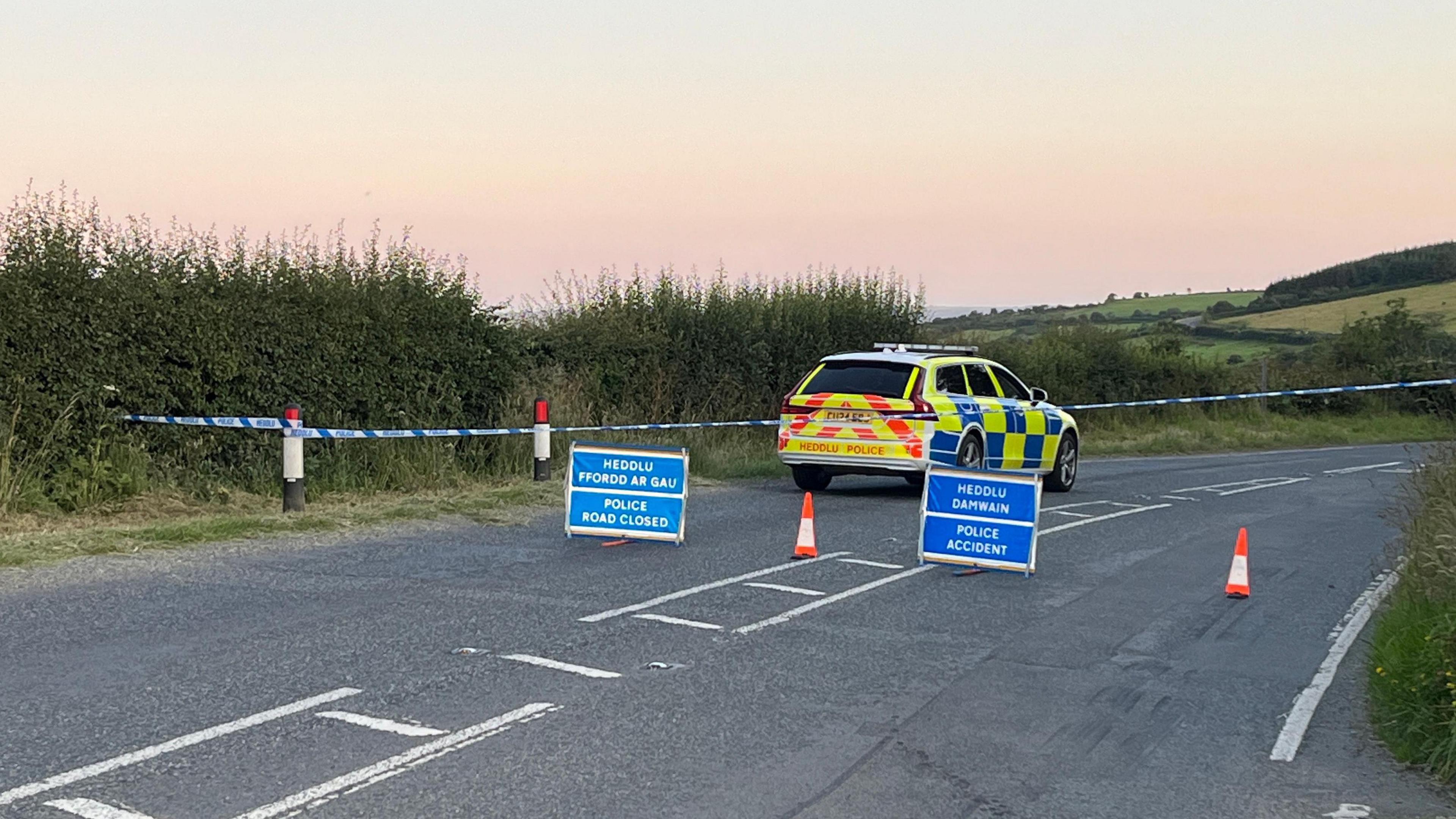 Police car stationary on road which has been cordoned off with blue and white police tape. Blue signs have been put up on the road saying Police Accident and Police Road Closed in English and in Welsh alongside orange and white cones. There is a bend in the road and hills and fields are visible behind.