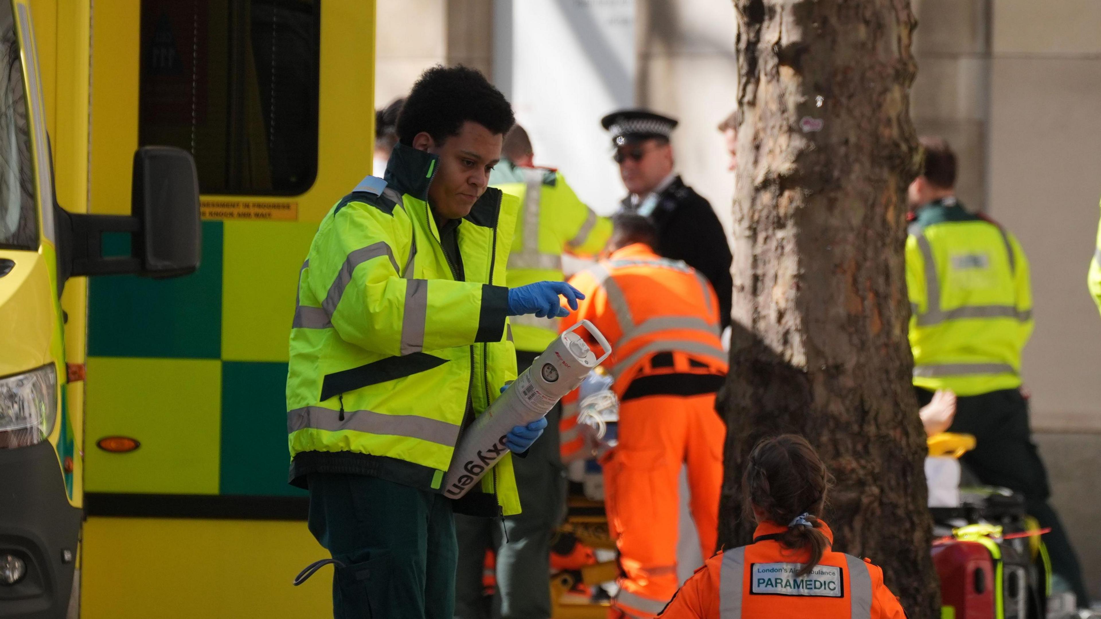 A paramedic wearing a yellow fluorescent jacket holding an oxygen container beside a yellow and green ambulance. Two paramedics in fluorescent orange jackets, and a uniformed police officer, can be seen on the right in the distance