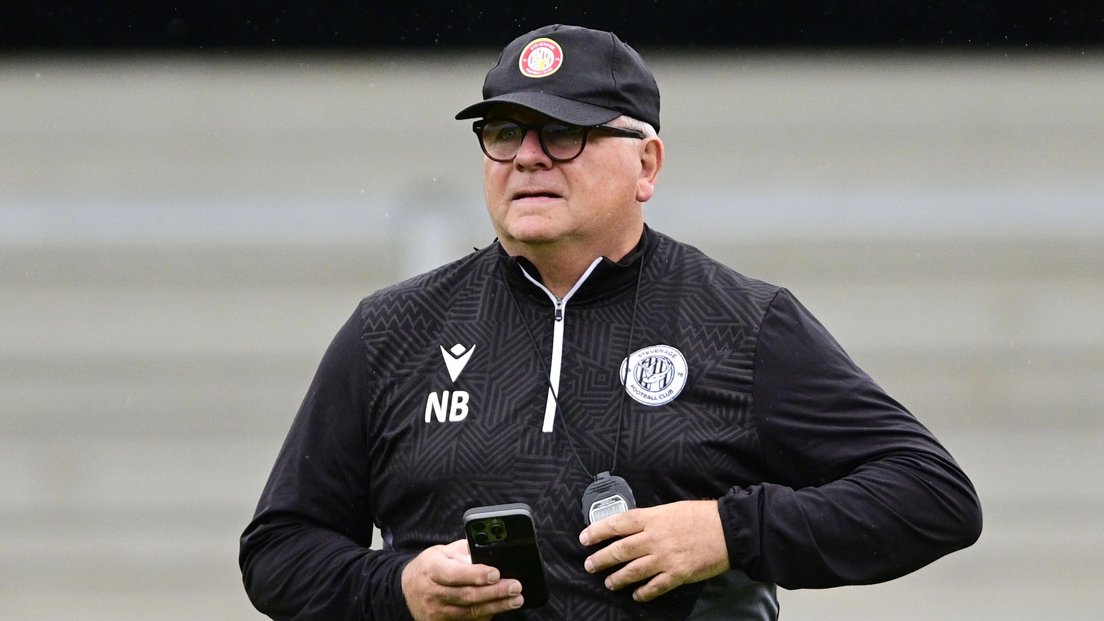 Neil Banfield wearing a black top with a Stevenage badge and NB written on it and a black cap. He is holding a phone in his hand