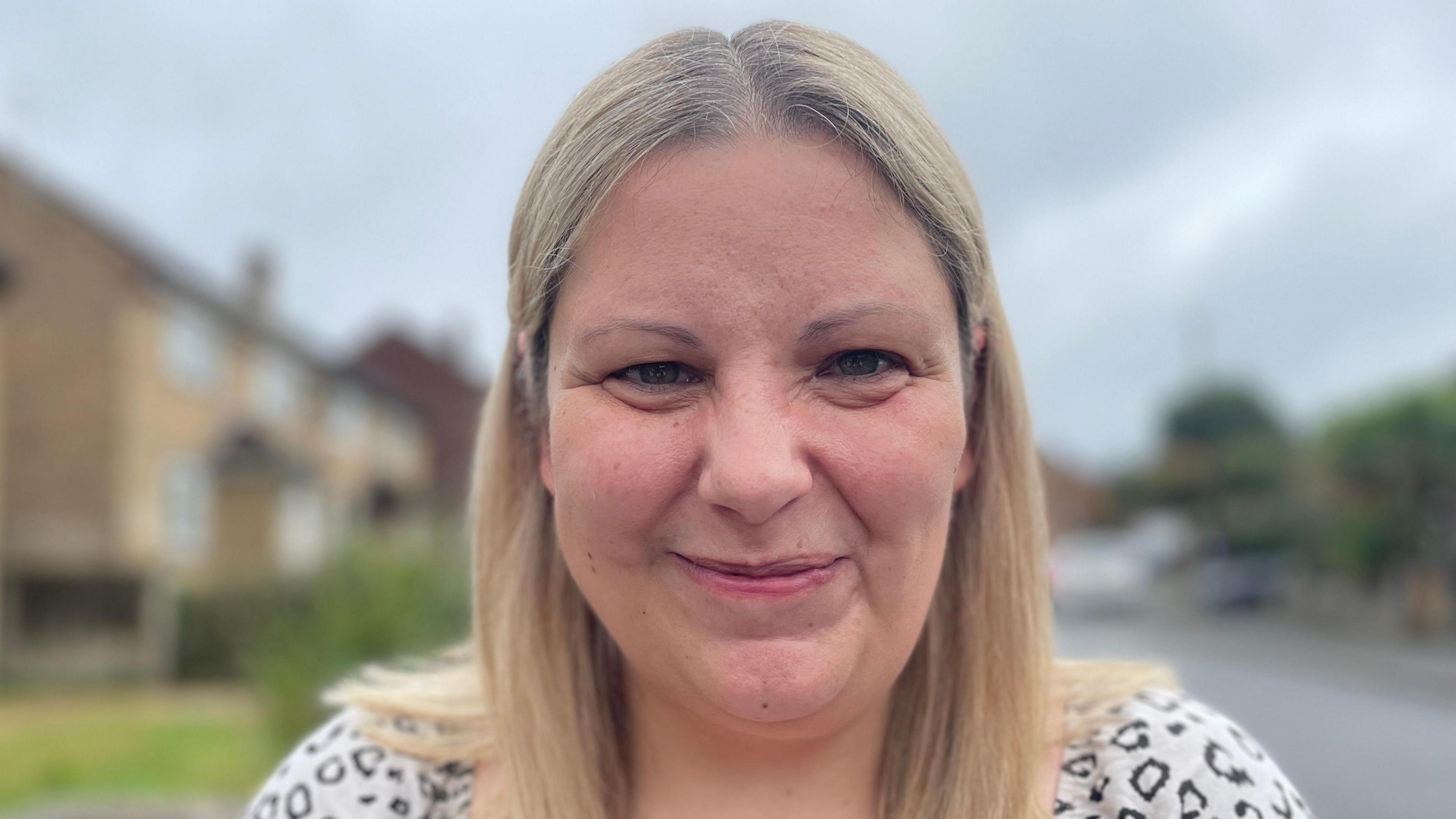 Nicola is smiling, she has brown eyes and freckles dappled across the right side of her cheek. She has a long blonde bob and is standing in front of a road which has been blurred.