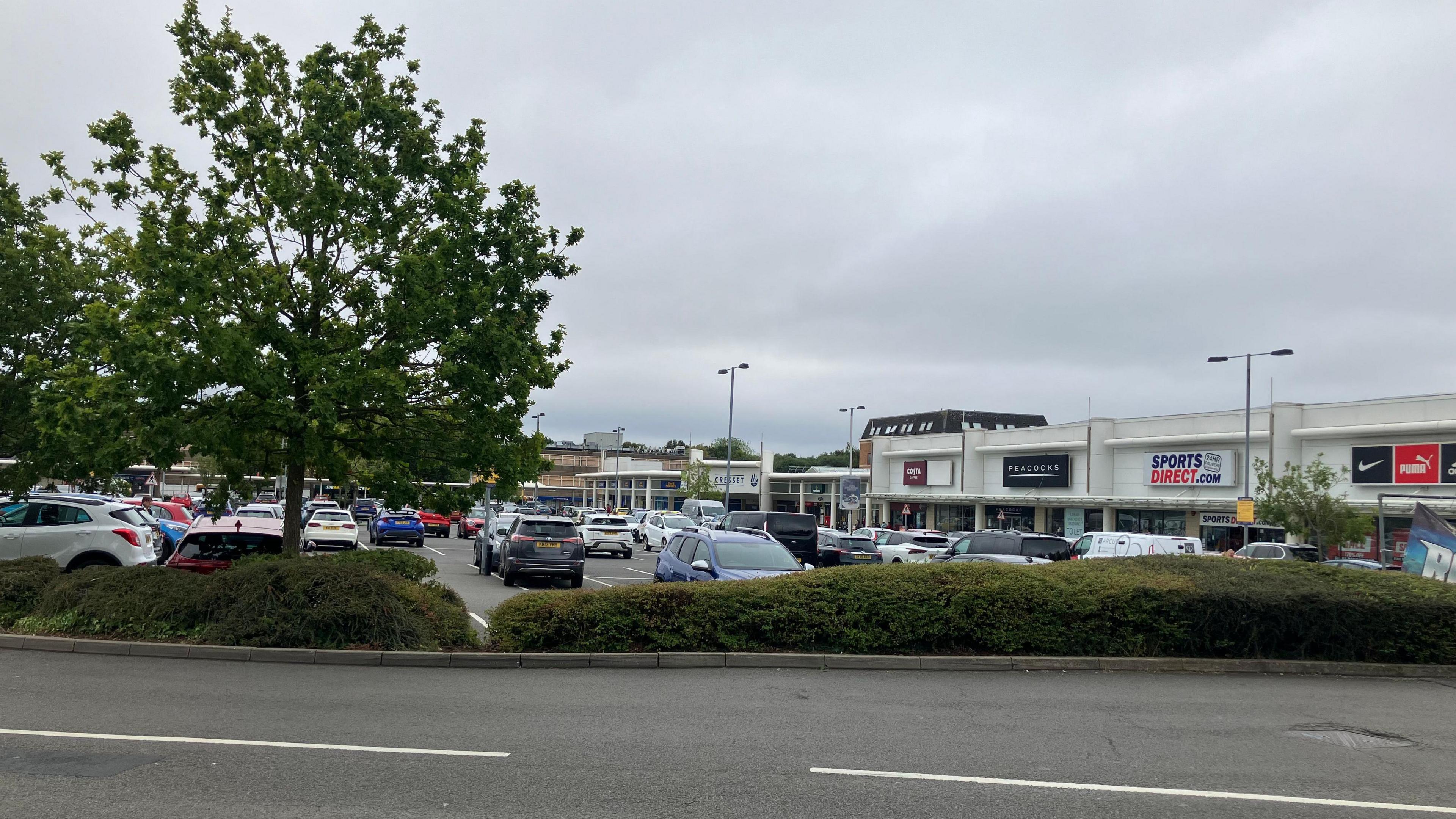 Outdoor area and car park of  the shopping centre with a white building- with names of different businesses including Sports Direct, a maroon Costa board and a black Peacocks board.