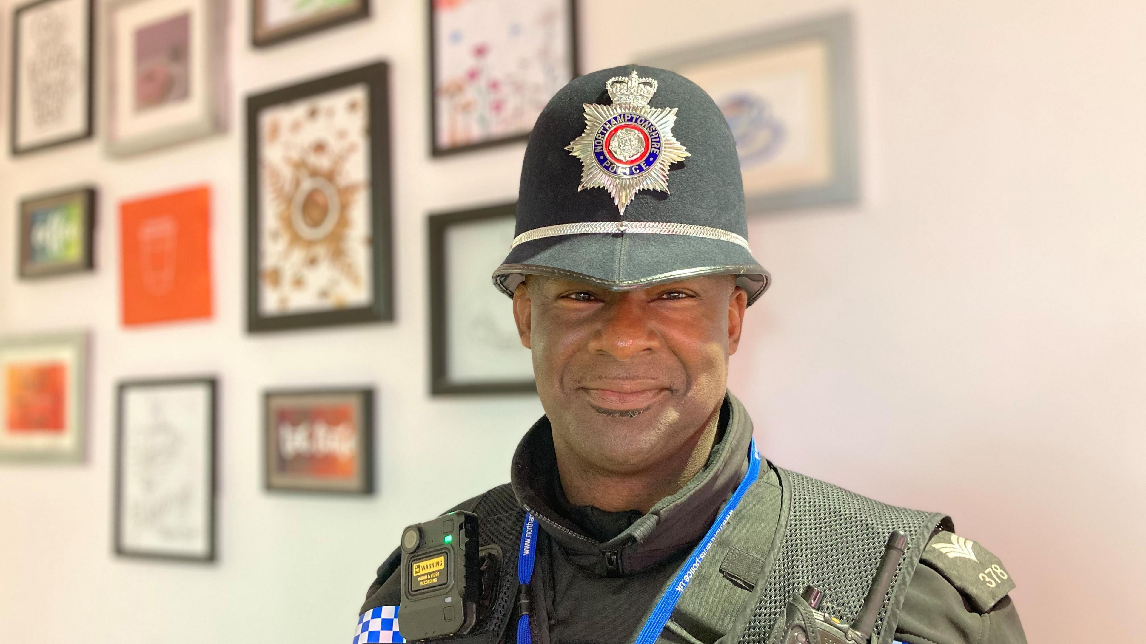 Sgt Williams wearing police uniform with helmet.  He has a blue lanyard around his neck and a radio on his right shoulder.  There is a wall behind him on which various framed pictures are hanging.