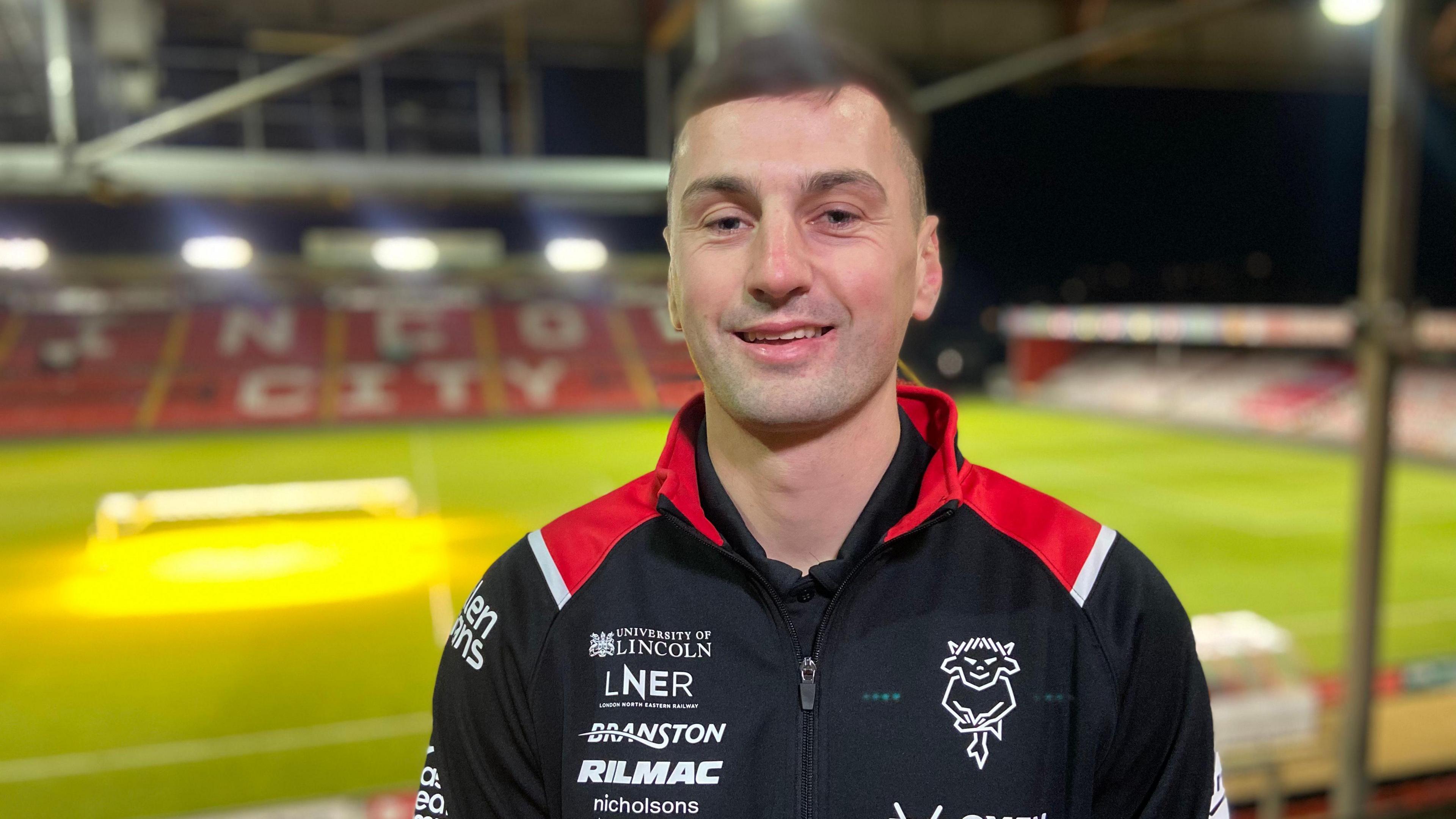 Footballer Paudie O'Connor stands in the Lincoln City FC stadium. He is smiling and short brown hair. He is wearing a black club tracksuit top with red trim which covered in sponsor logos and the football club badge. The green football pitch is visible in the background alongside the red and white-coloured seats.