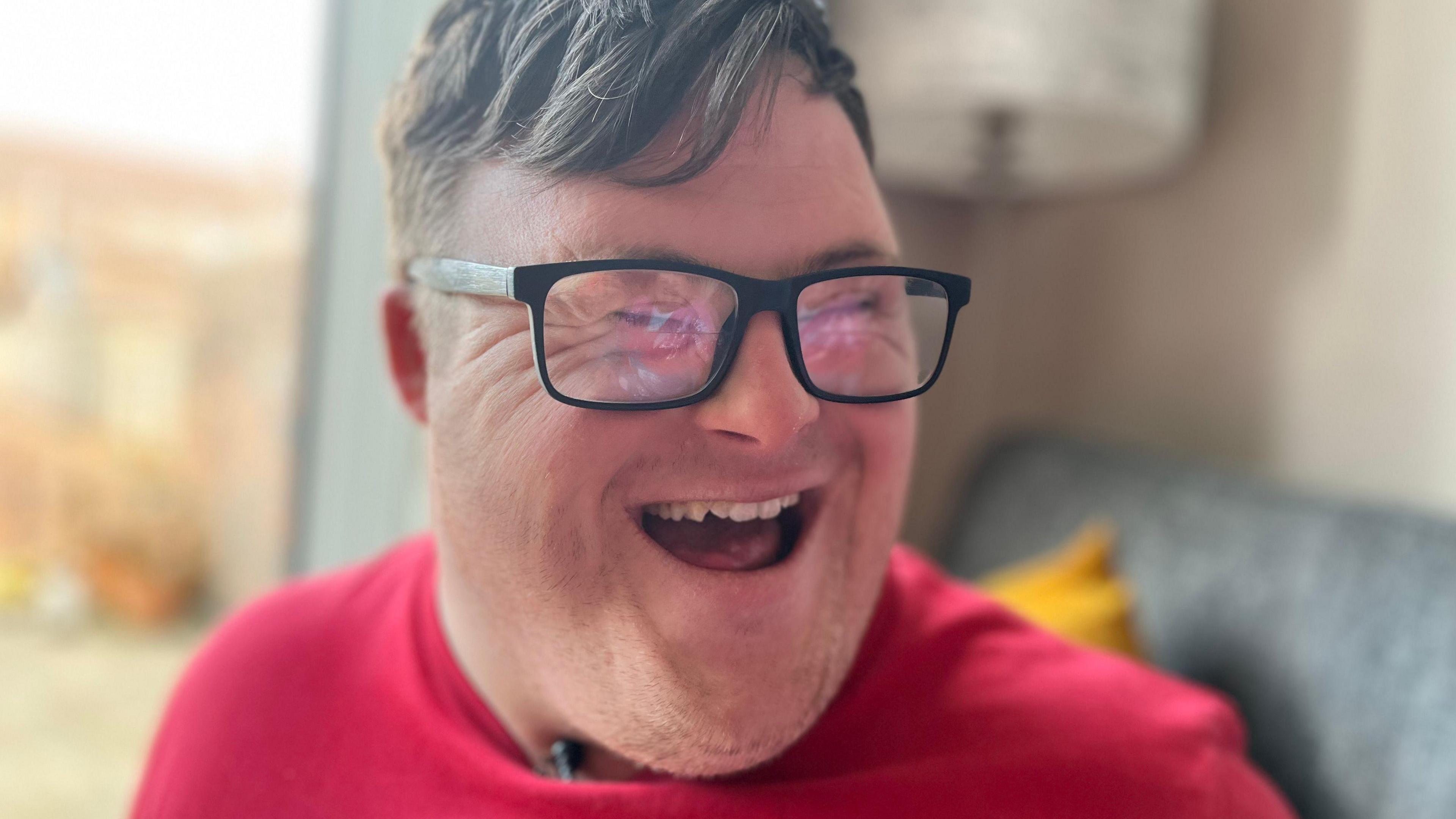 A close-up head shot of Joel laughing. Joel has Down Syndrome, has short brown hair and is wearing a red, green and yellow t-shift and dark-rimmed glasses. He is sitting on a grey sofa. 