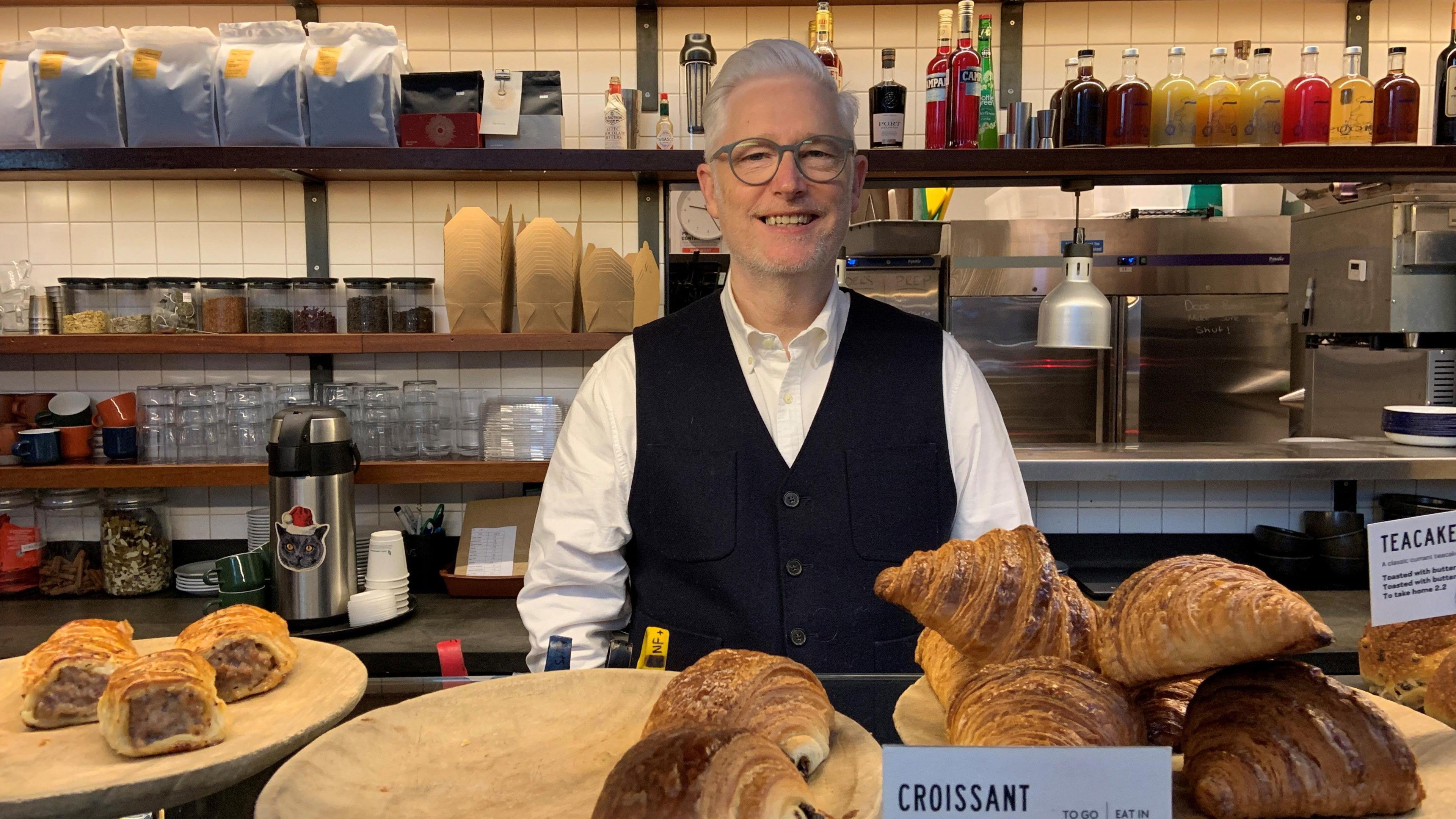 A man has white hair in a quiff and is wearing glasses, a white shirt and black waistcoat. He is standing behind a counter with several croissants, in the background are shelves with coffee bags, a coffee machine, glasses and bottles