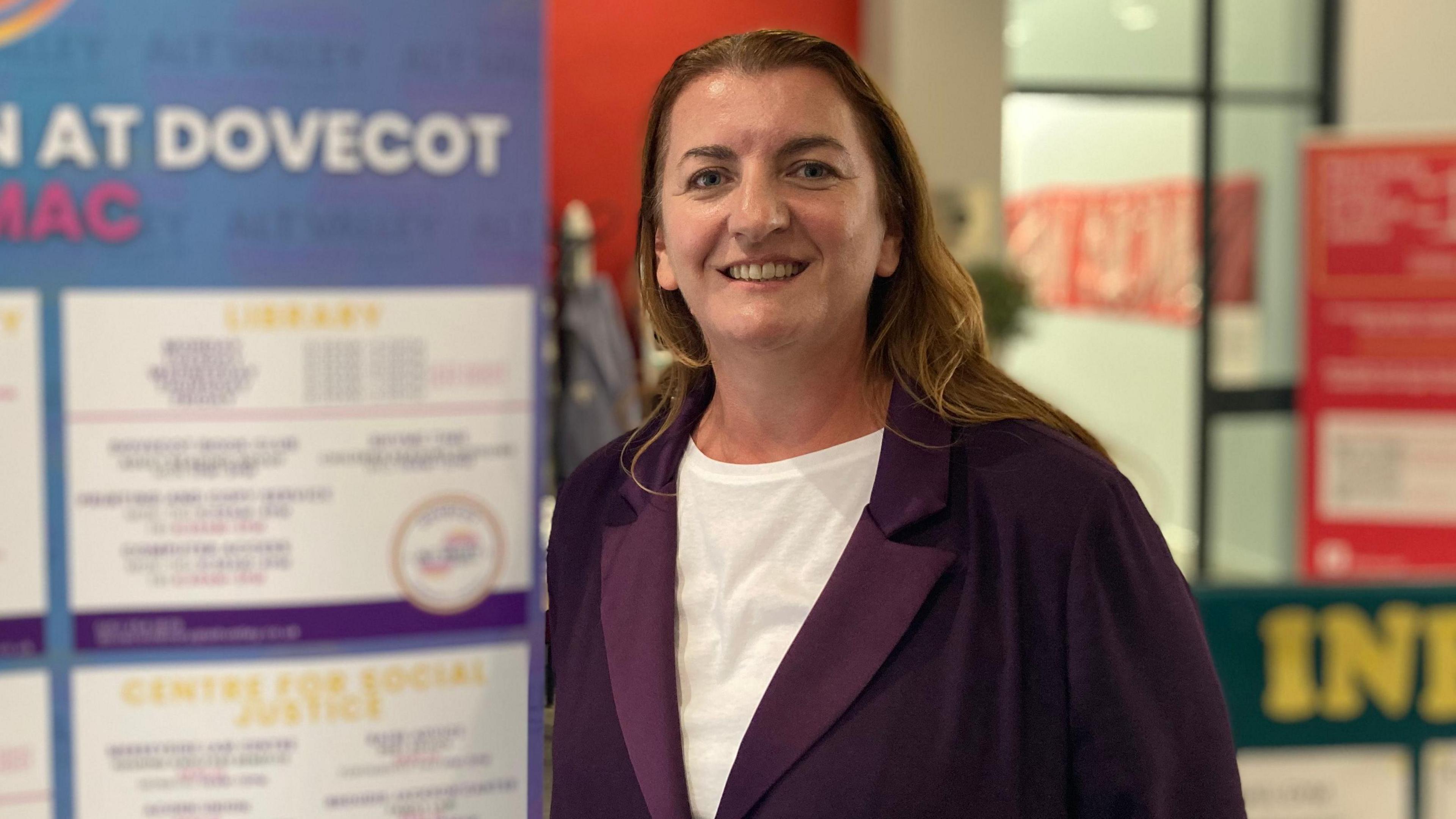 A woman stands smiling in front of a blurred background. She has auburn shoulder length hair, and is wearing a purple jacket and white t-shirt.