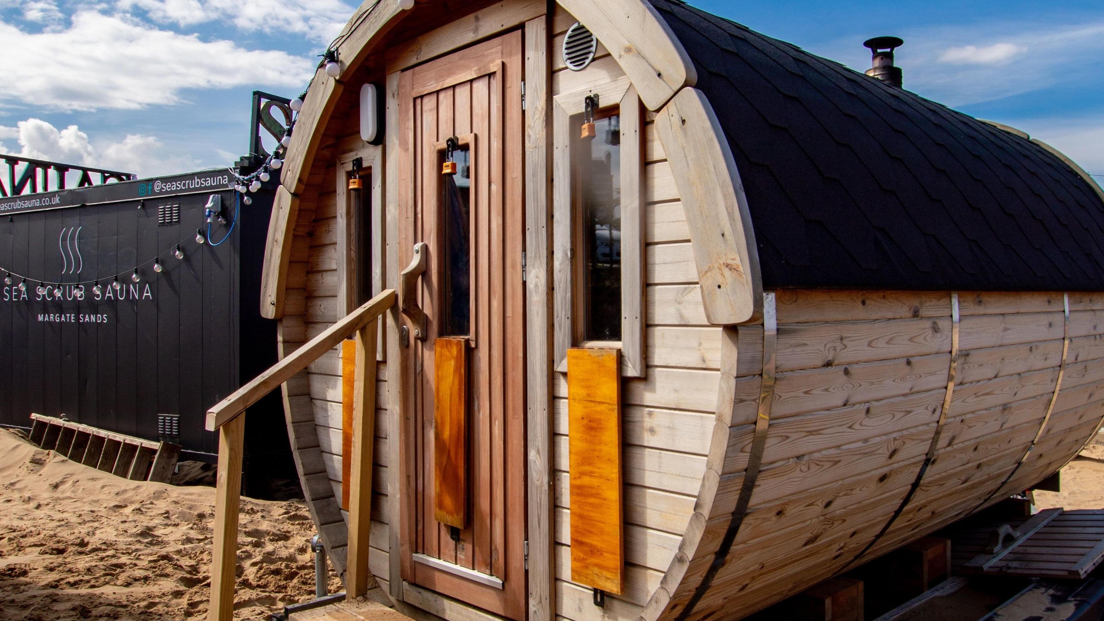 One of the wood saunas at the Sea Scrub Sauna on Royal Crescent Promenade in Margate; this circular sauna is made out of wood and has a sauna heater flue at the back for smoke to exit. At the front there is a long rectangular door to enter, flanked by two smaller rectangular windows. To the left there is a bannister to walk to the entrance.