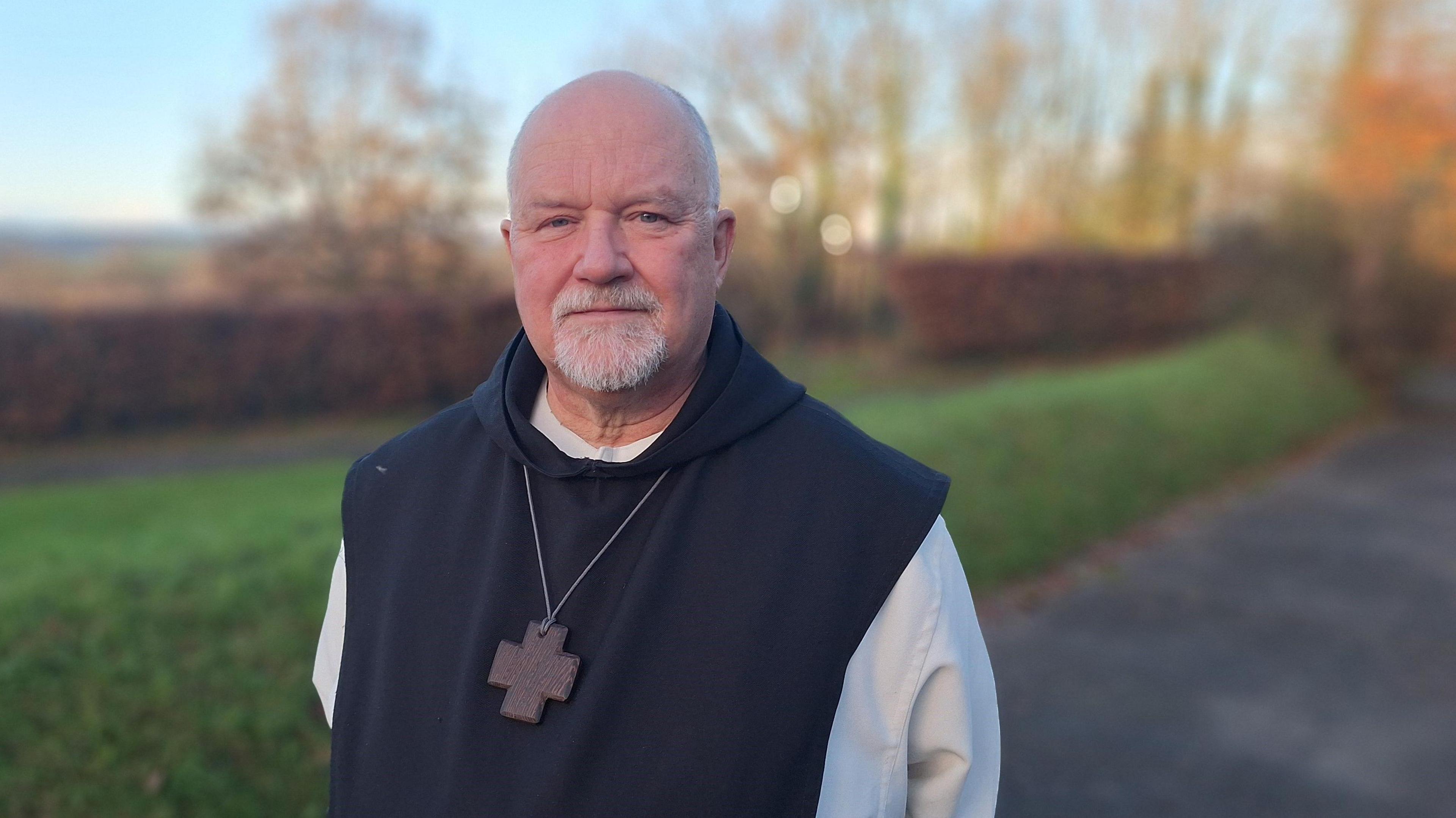 Father Jan Rossey stands in a traditional monk's habit - white robe covered in a black hooded robe with a cross on a long chain around his neck. His hands are behind his back and he's looking into camera, smiling slightly. He is bald with a white goatie beard.