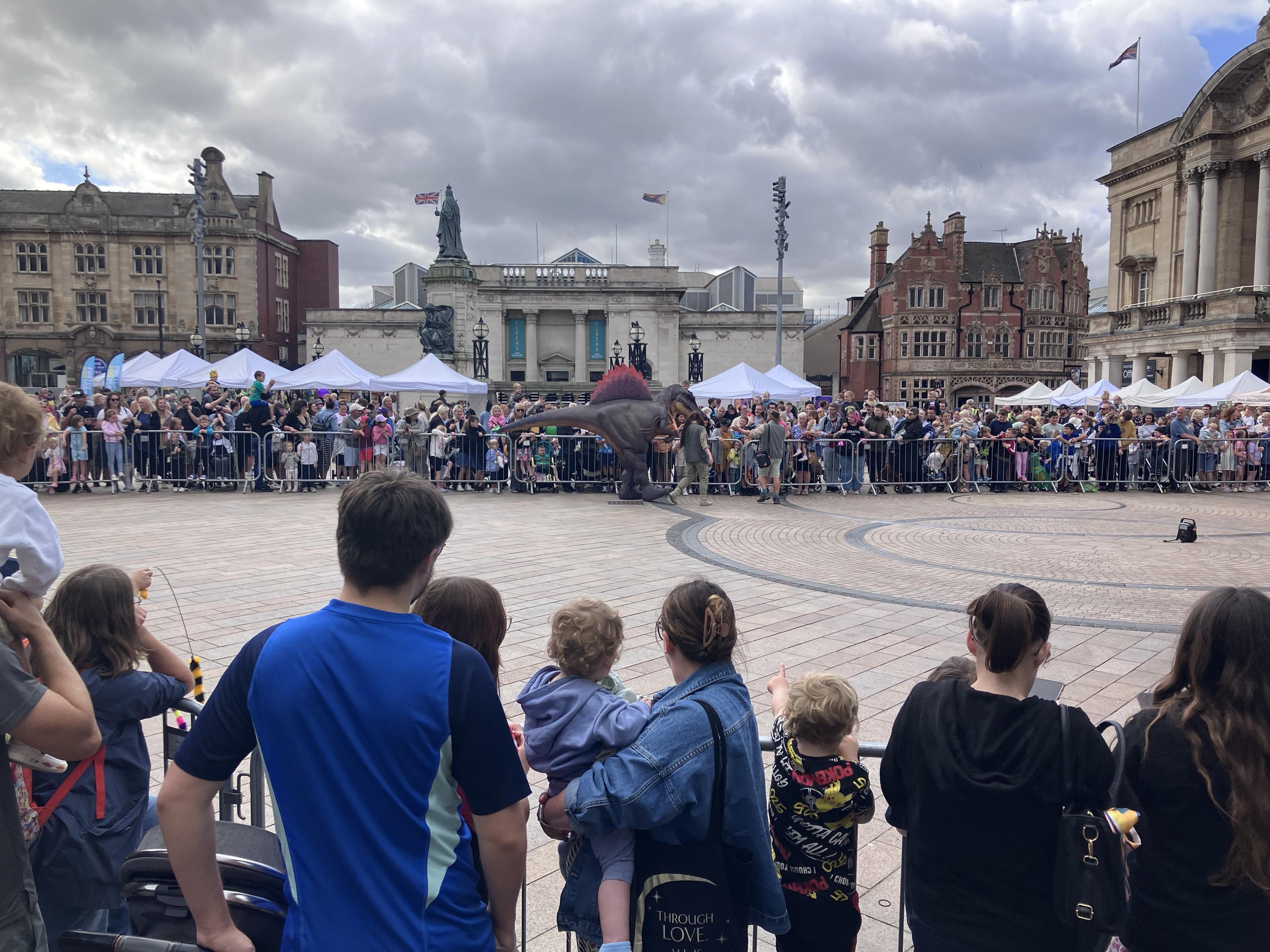 A wide shot of Queen Victoria Square. You can see people on all sides watching the dinosaur show. A person in a spinosaurus costume stands in the middle distance.