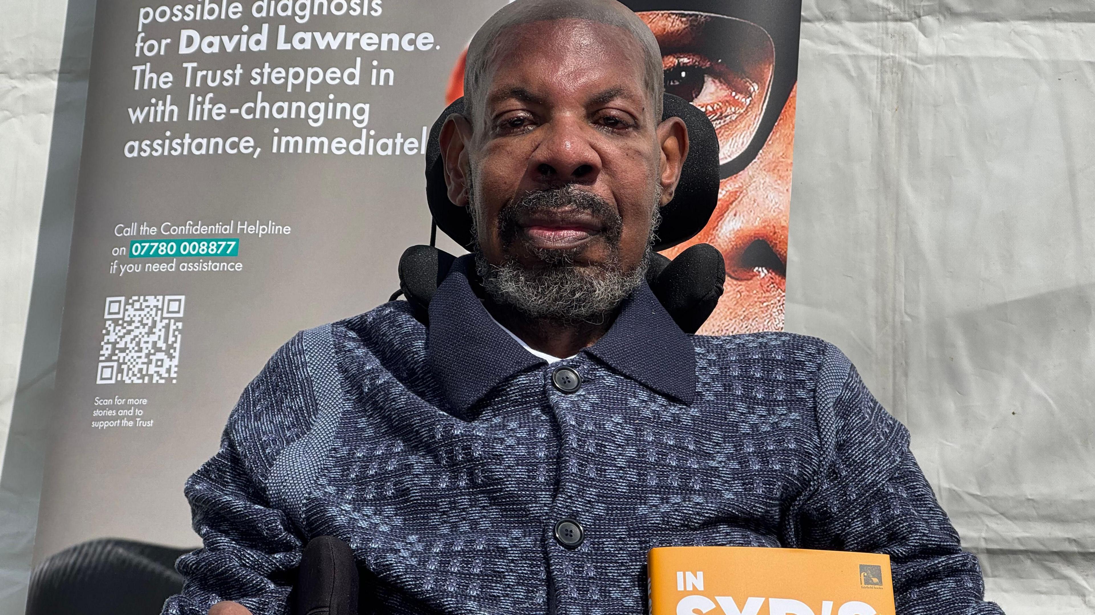 Syd sitting in a wheelchair with a dark polo shirt on, short beard and hair, smiling at the camera. There is a poster behind him and he is outside by a white tent.