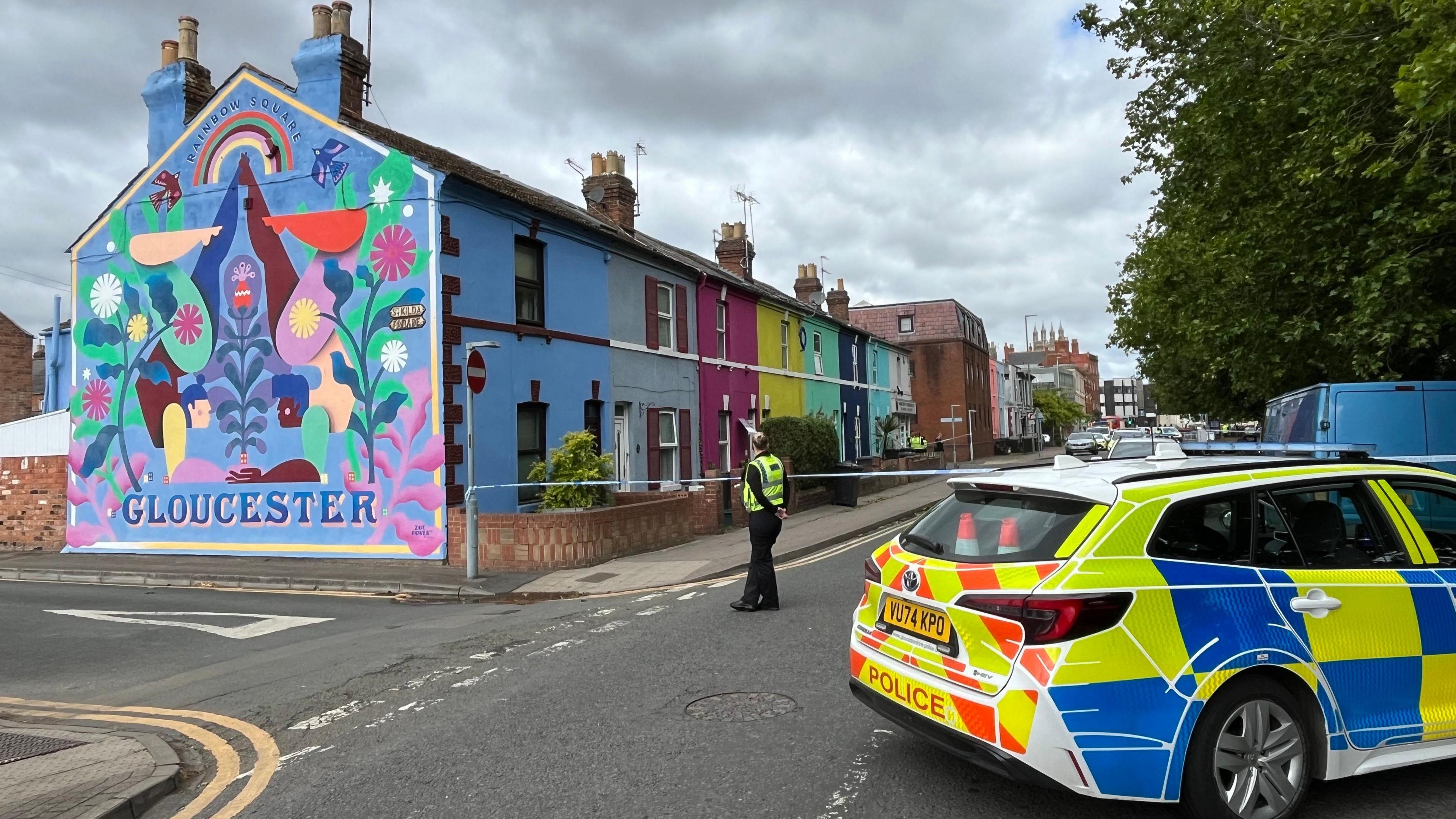 A police car is parked across a road in a residential city centre street. There is a female police officer standing on the road, with her hands behind her back as she looks at a row of colourful houses. There is police tape across the road. The end terrace house has a painted mural that reads "Gloucester" on it, along with two people holding hands as they do handstands among floral imagery and a rainbow.