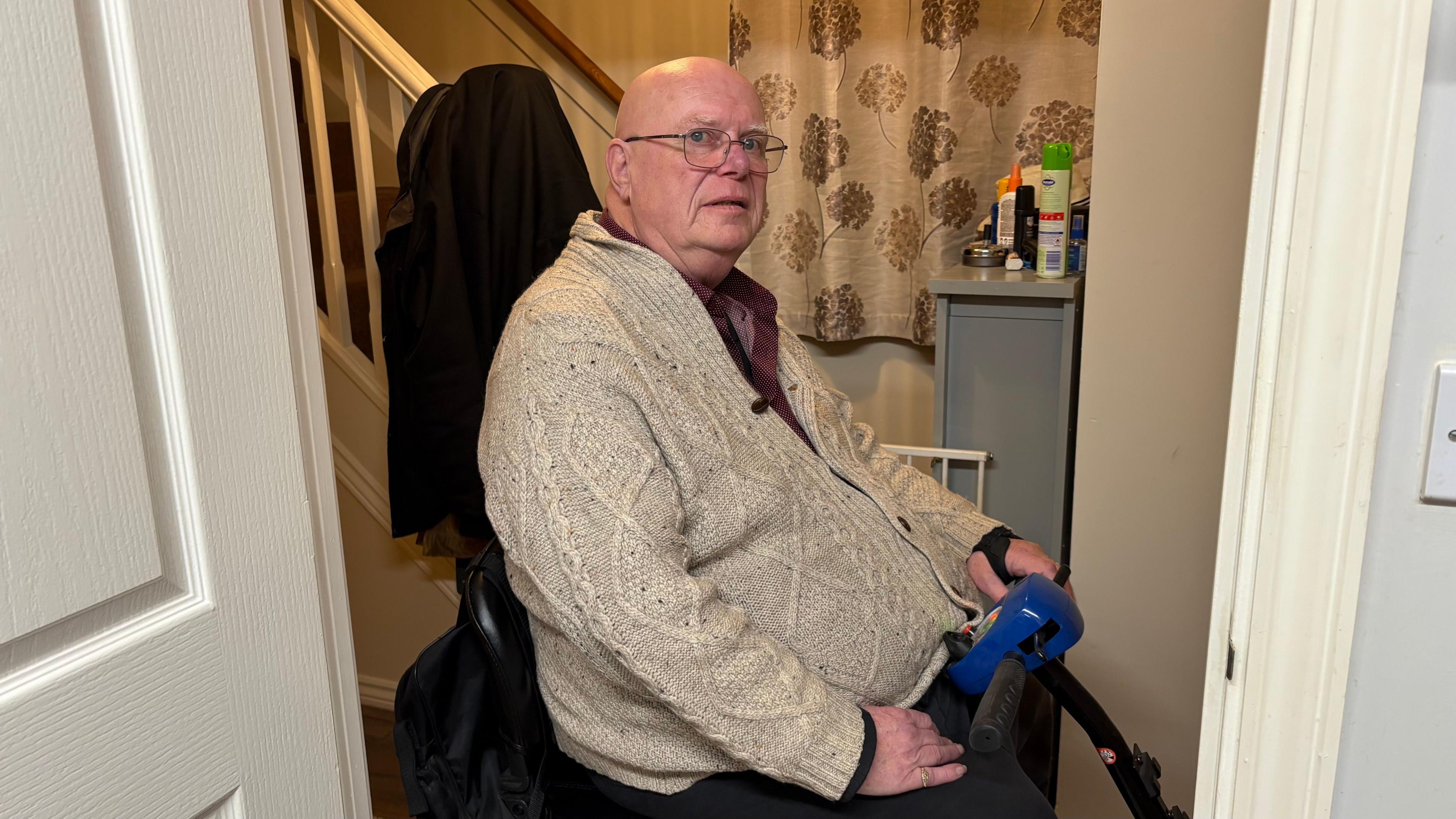 Andy Watson with short hair and glasses wearing a beige cardigan and sitting on a mobility scooter with a blue steering wheel. There are stairs and brown curtains behind him, as well as a grey chest of drawers on which several aerosol cans are resting.