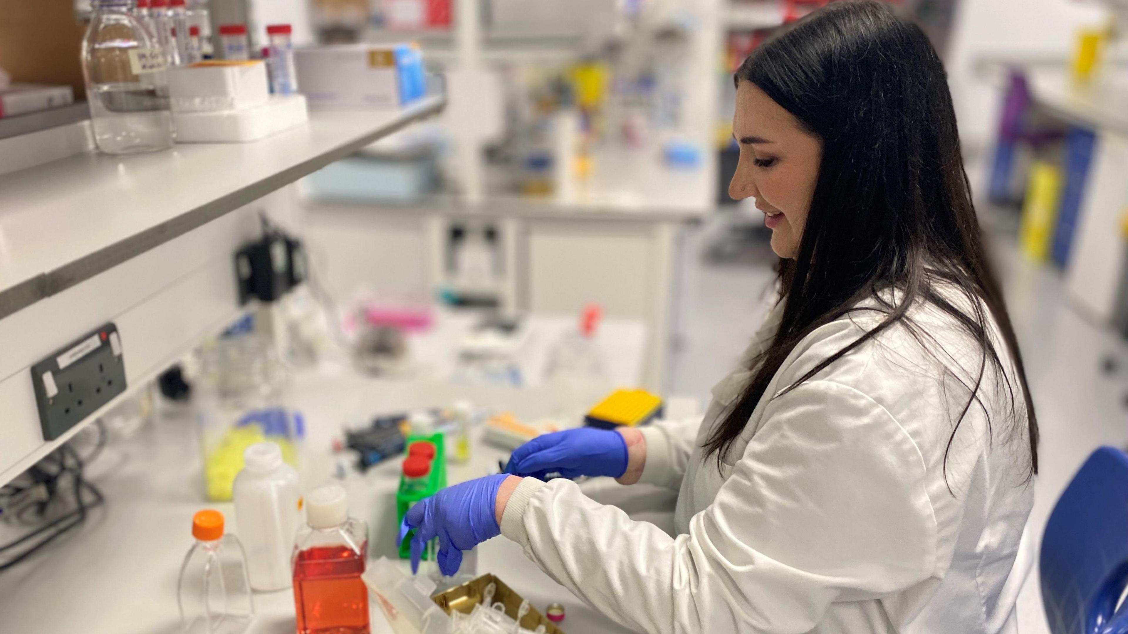 Becca Heritage in a science lab. She is wearing blue gloves and a white lab coat. On the table in front of her are several bottles with coloured fluids in them. Shelves above her workstation also have vials and bottles on them.