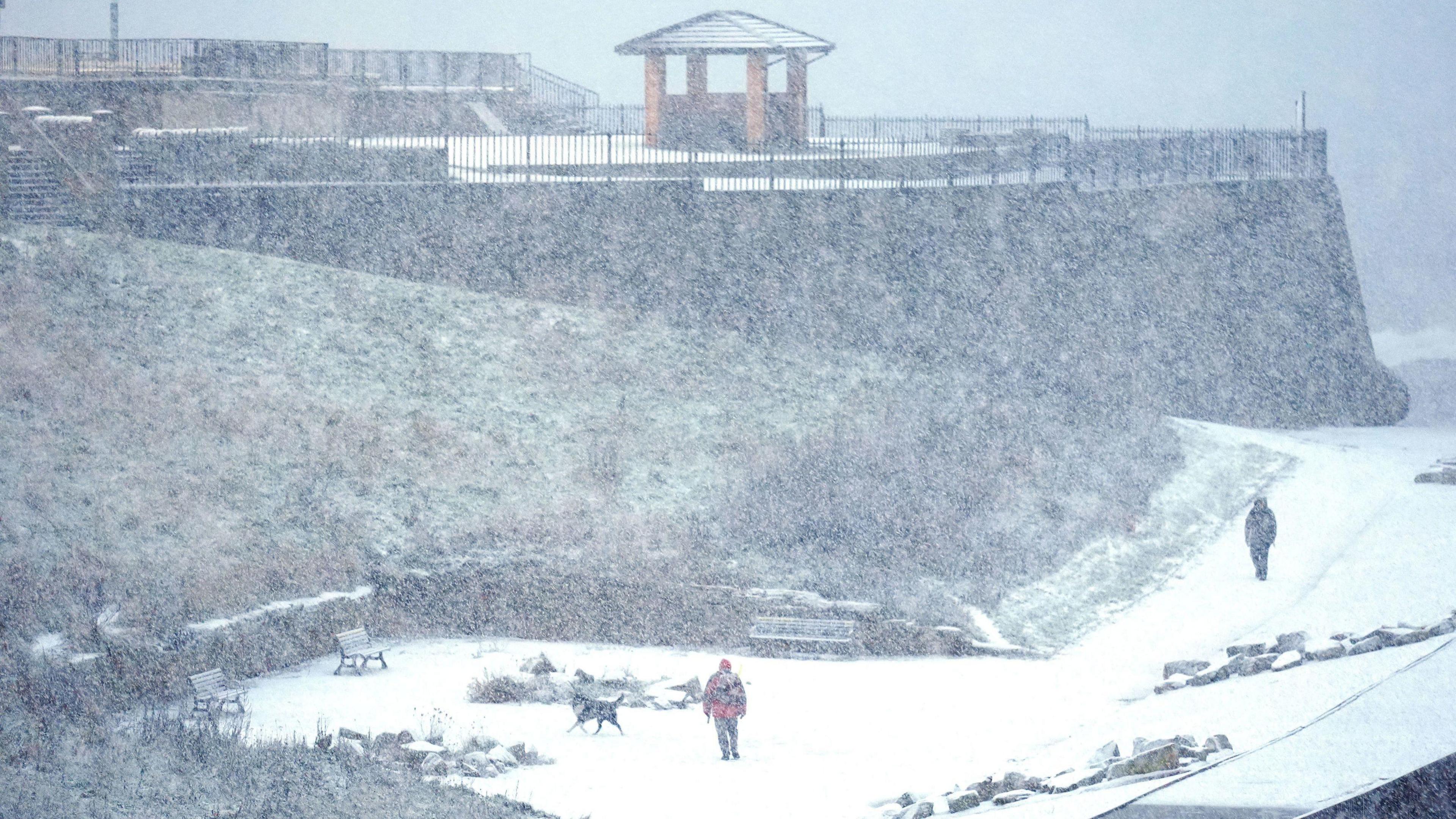 People walking in the snowy conditions in Cullercoats on North Tyneside. Snow is falling over the cliff edge onto the promenade below.