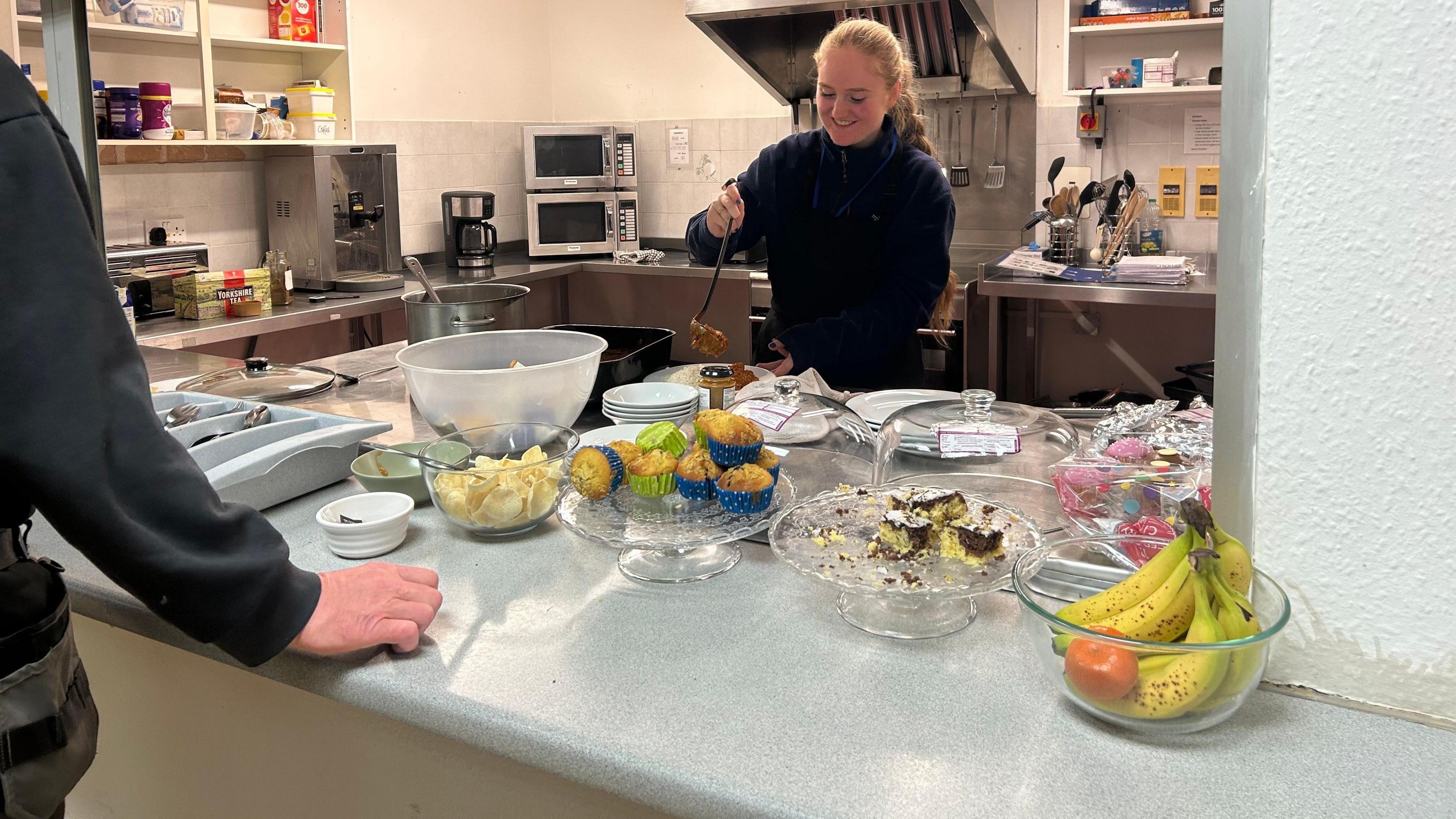 A smiling volunteer works in a kitchen, ladling out food. She is looking down as she plates up a dish and is wearing a dark top. There is food in front of her and a kitchen behind her. A man's arm can be seen to the left. 