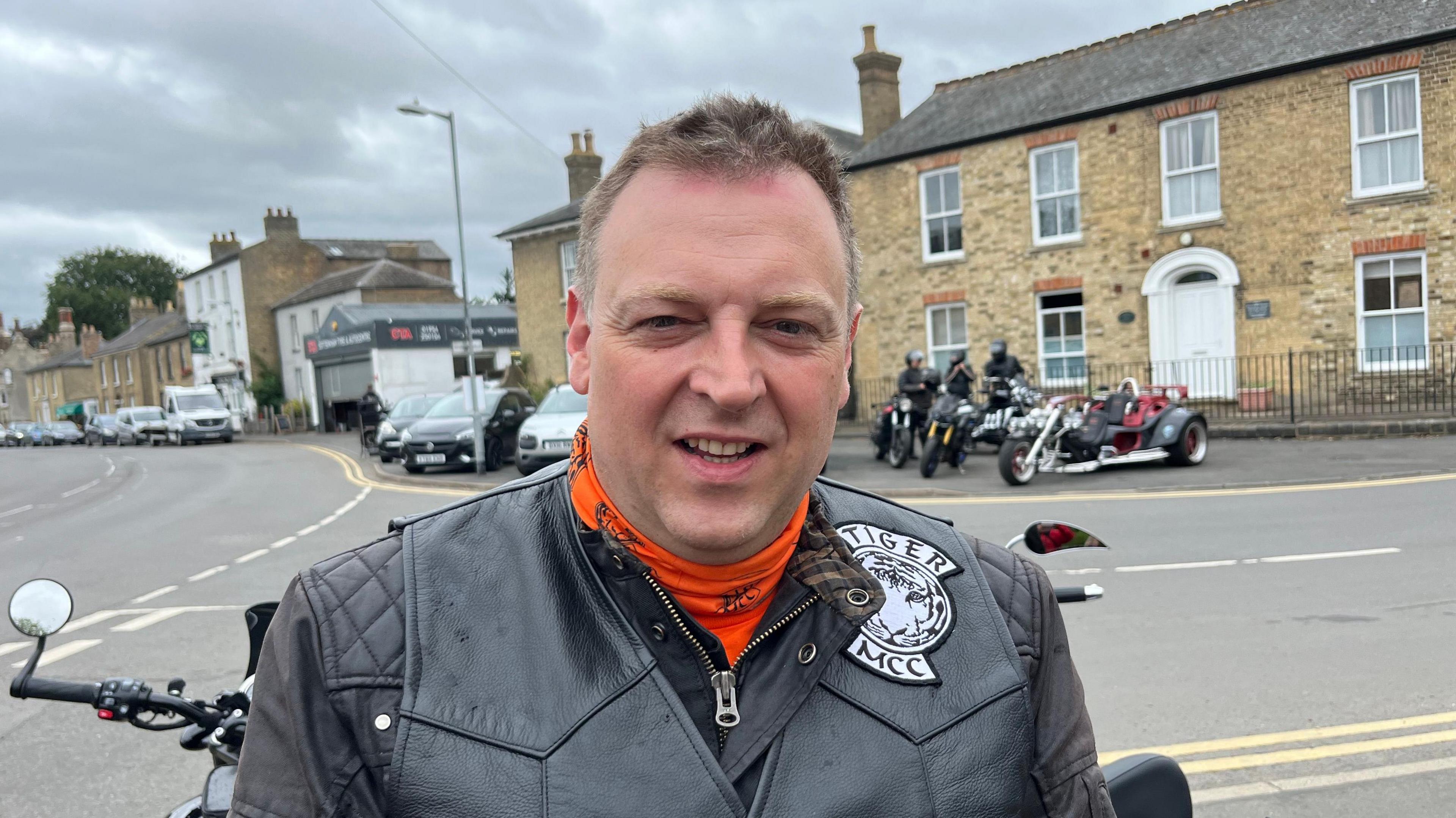 Sam Harris is looking at the camera. he has short fair har and is wearing a black leather jacket and an orange top. There are motorcycles and riders on the side of the road behind him