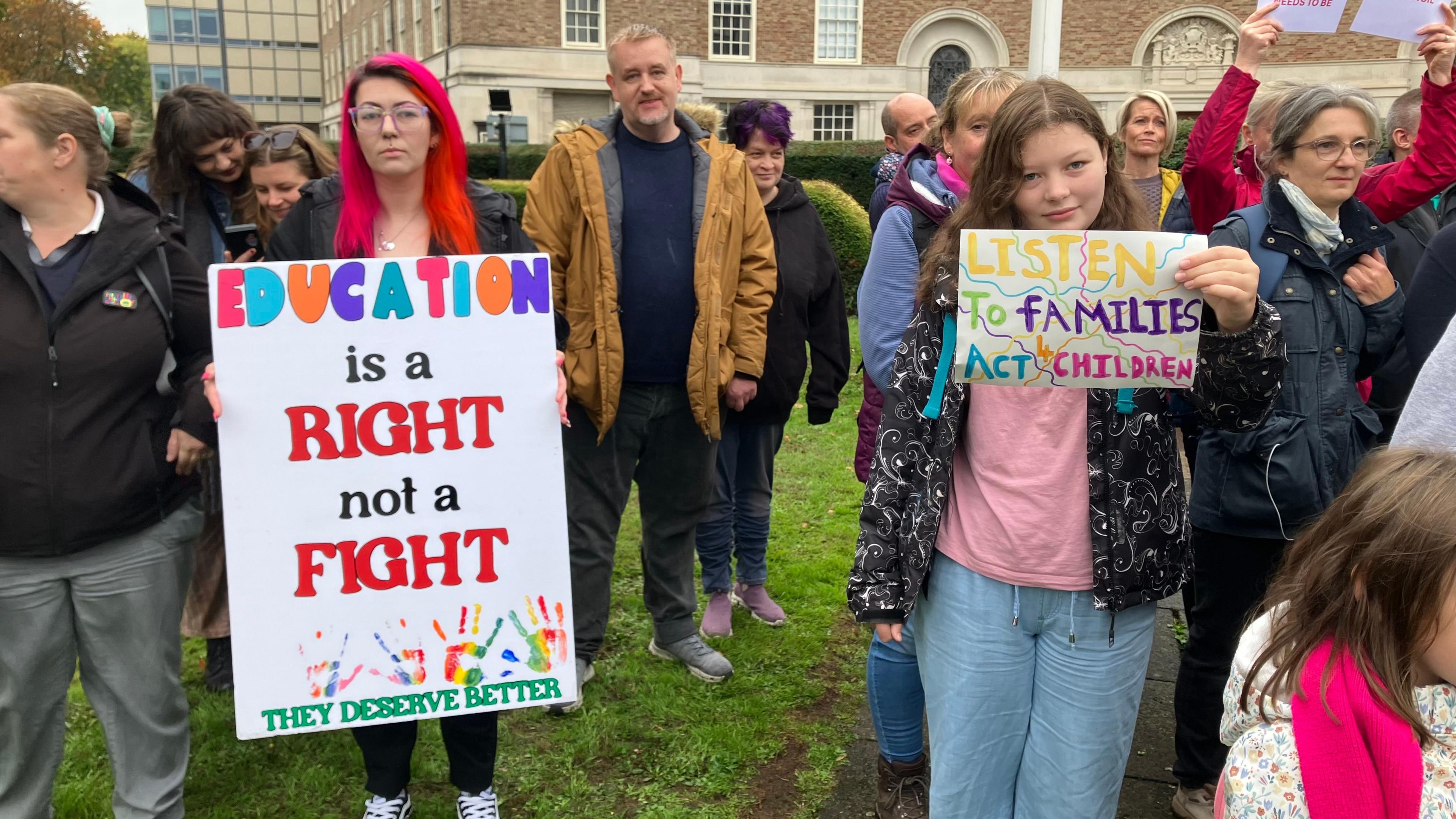 A group of people standing on grass outside a municipal building holding placards reading things like 'education is a right not a fight'