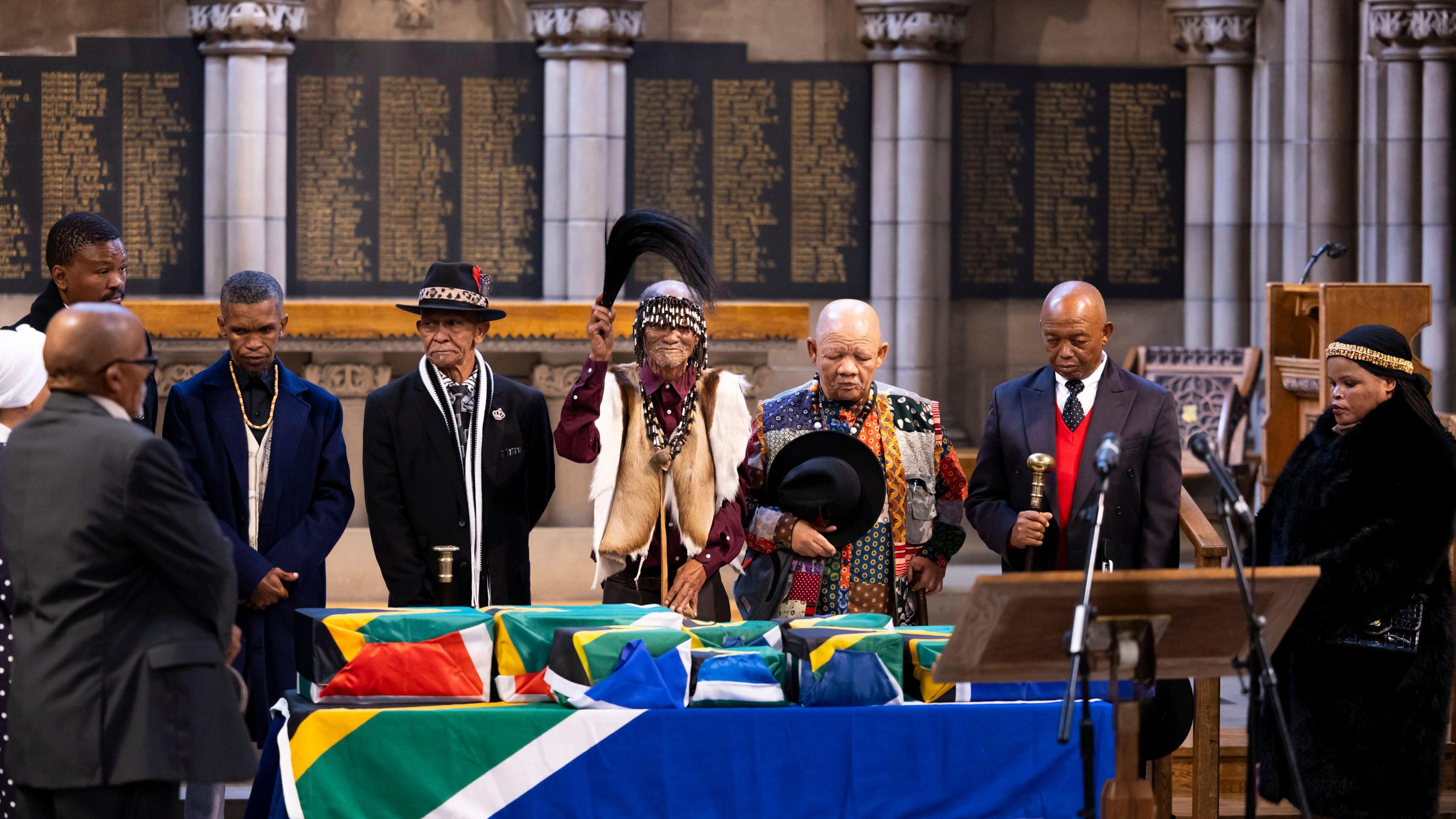 Several people standing over coffins draped with South African flags, containing the remains of their ancestors dating back over 100 years. Many of those standing have their heads bowed.