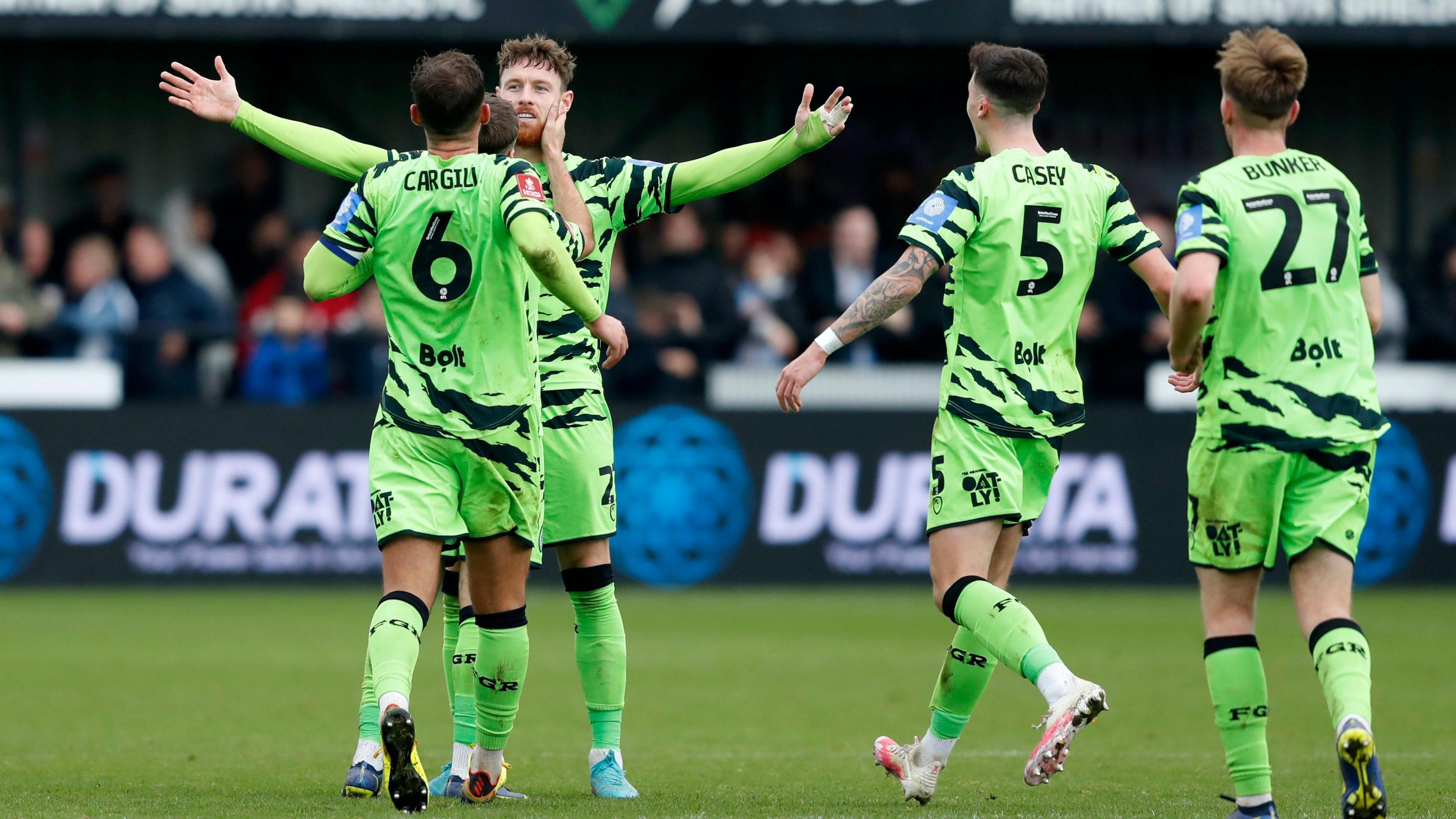 Four teammates of the Forest Green Rovers football club celebrating on the pitch after scoring a goal. They are wearing their old football kit, which is fluorescent green with jagged stripes and matching socks.
