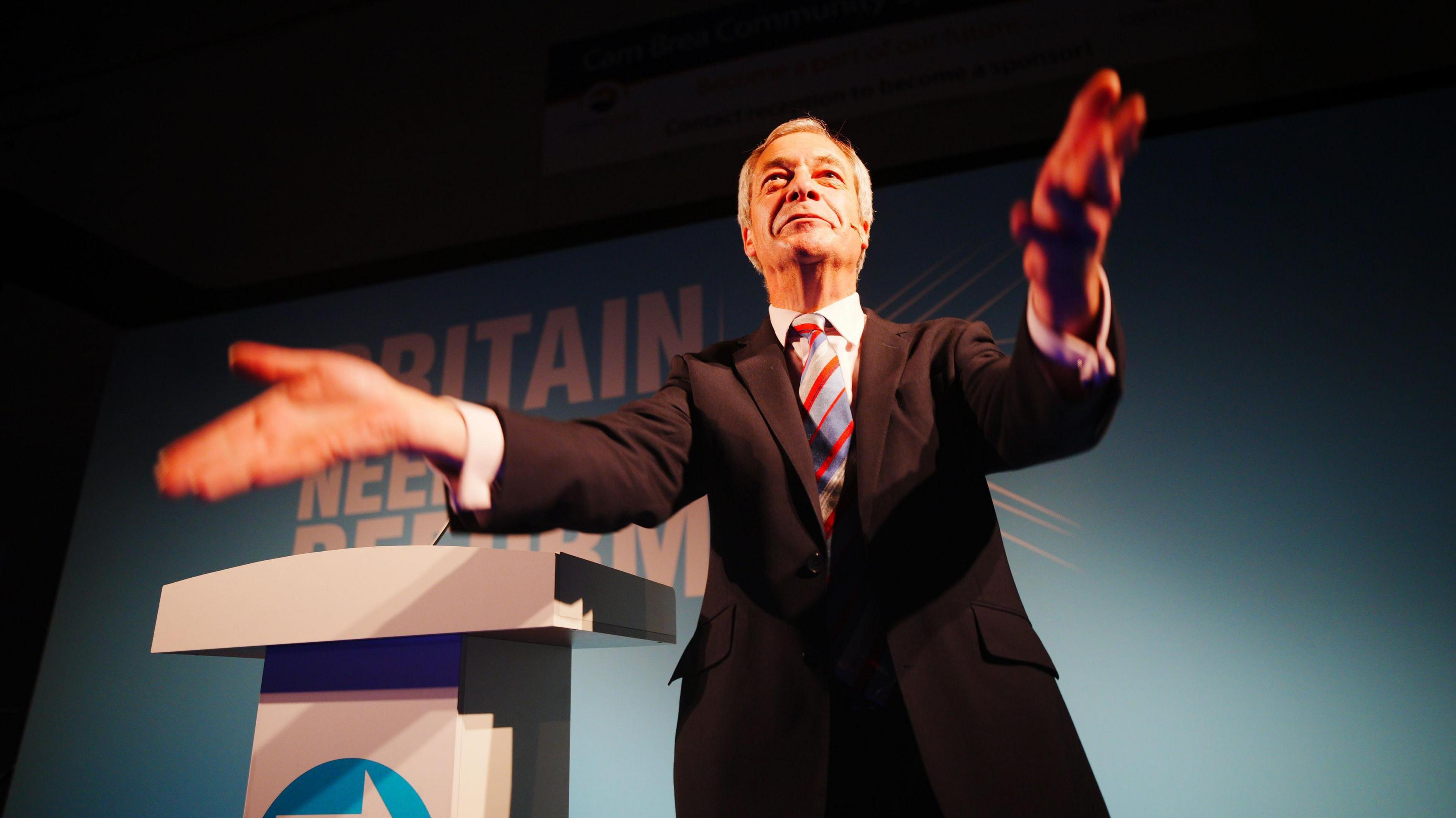 Nigel Farage in a conference podium, gesticulating. He is smiling while wearing a dark suit and a striped tie