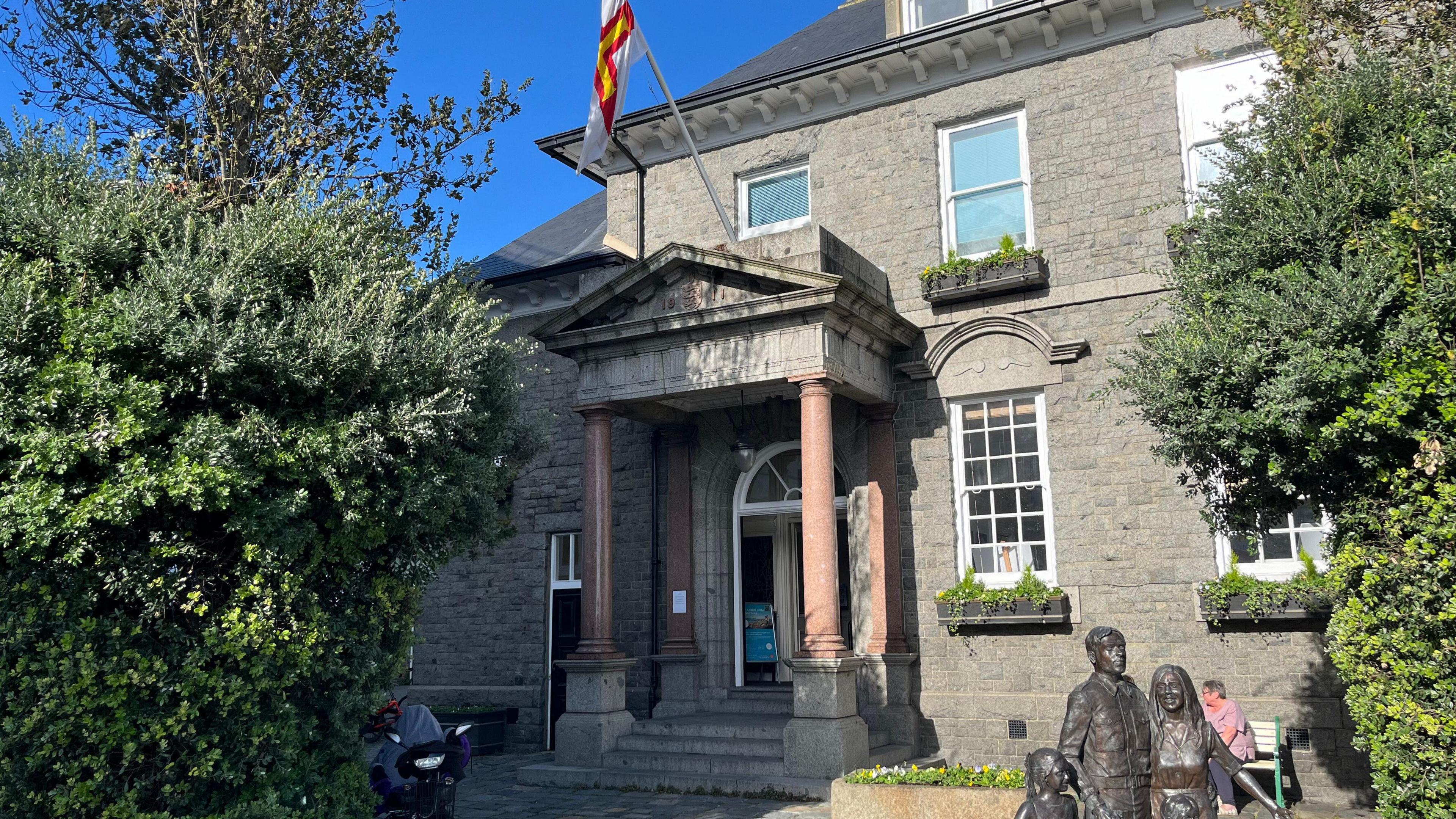 The Guernsey Information Centre. A grey building with coumns in the entrance with a statue of three bronze people in the bottom right. 