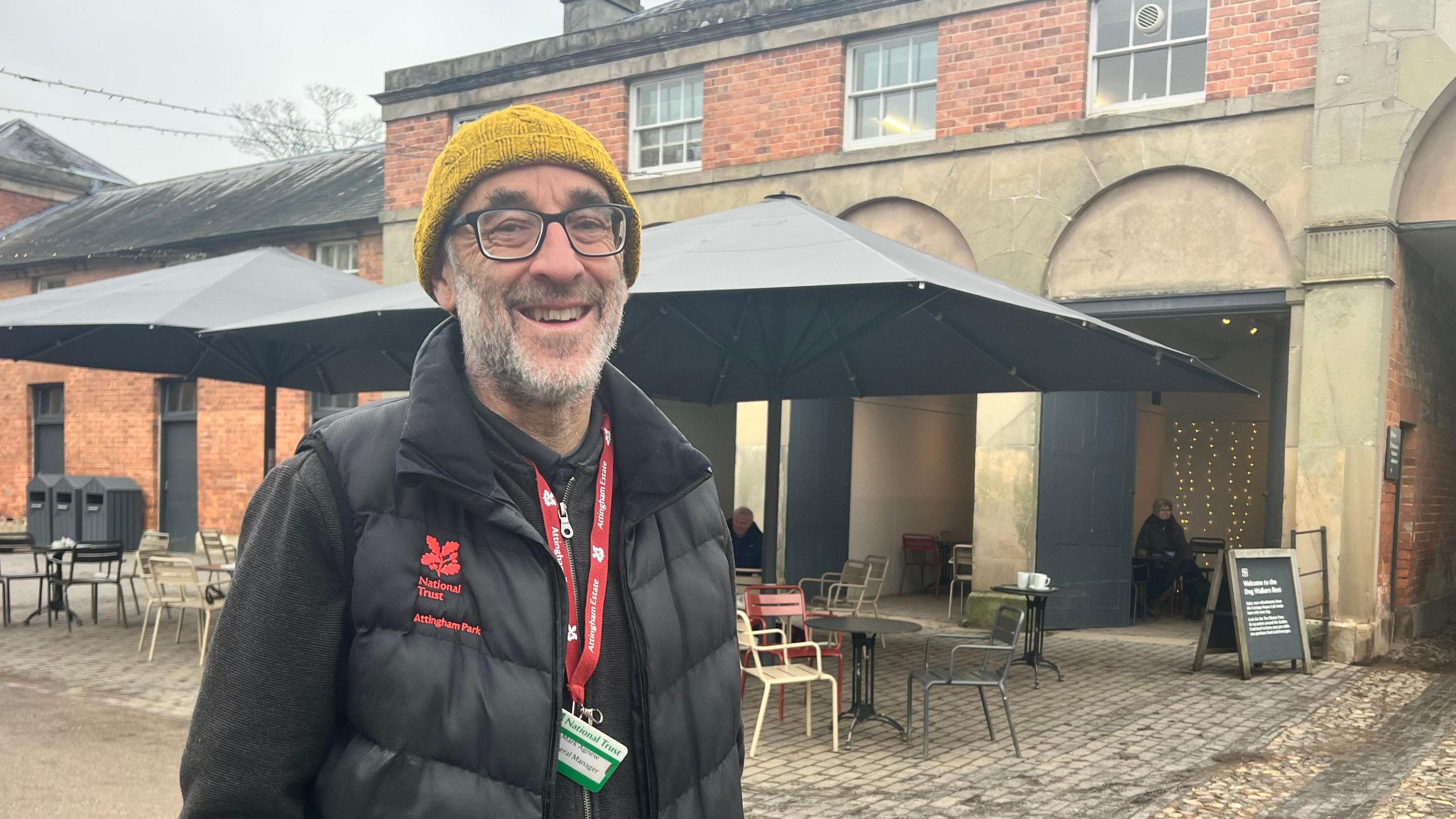 Mark looking into the camera and smiling. He's wearing a black fleece, a yellow beanie and a black gilet with the National Trust oak leaf logo and 'National Trust: Attingham Park' embroidered on it in red. He's also got a red lanyard and a green and white ID badge. He's stood in a courtyard of the estate, with chairs and tables behind him from the site's cafe. The buildings in the background are red brick, with Regency-style columns and arches. 