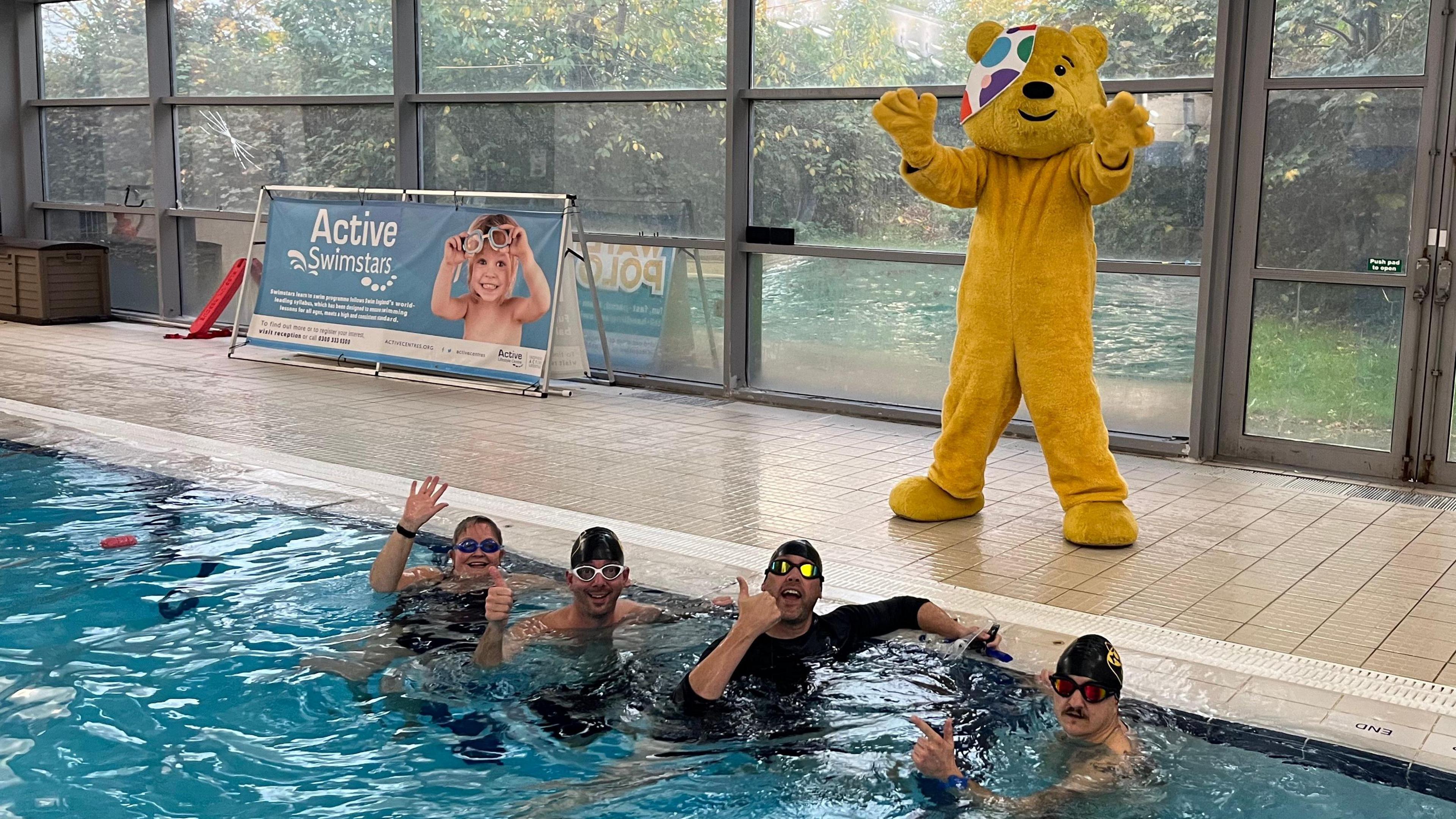 Four BBC Radio Bristol staff members in a swimming pool with Pudsey standing at the side of the pool waving at the camera with both hands