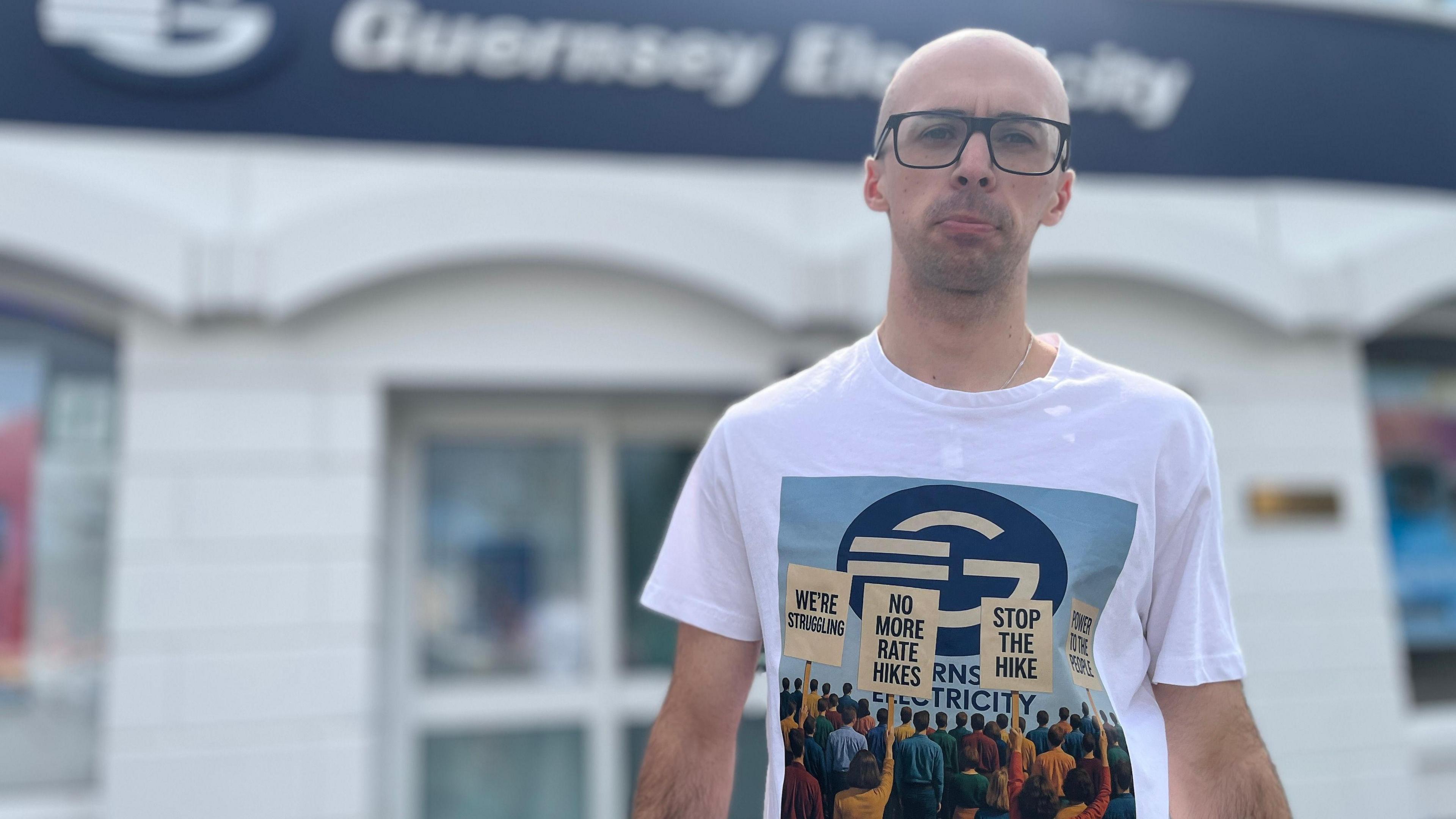 A bald man wearing glasses stands in front of Guernsey Electricity's head office. He is wearing a T-shirt with placards saying: "We're struggling! No more rate hikes, Stop the Hike and Power to the People."