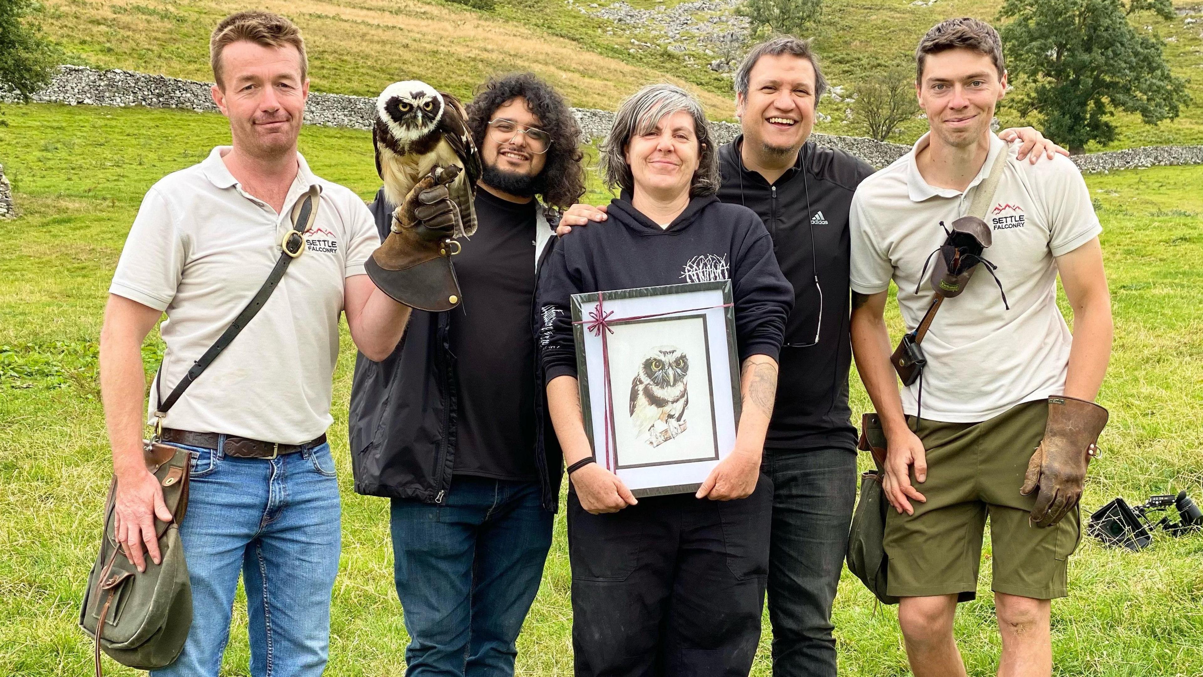 David Edwards and Kieren Bathers of Settle Falconry with the film crew and Elton the owl. The film crew wear all black, and David and Kieren wear light coloured polos. The group stands in a field in the Yorkshire Dales.