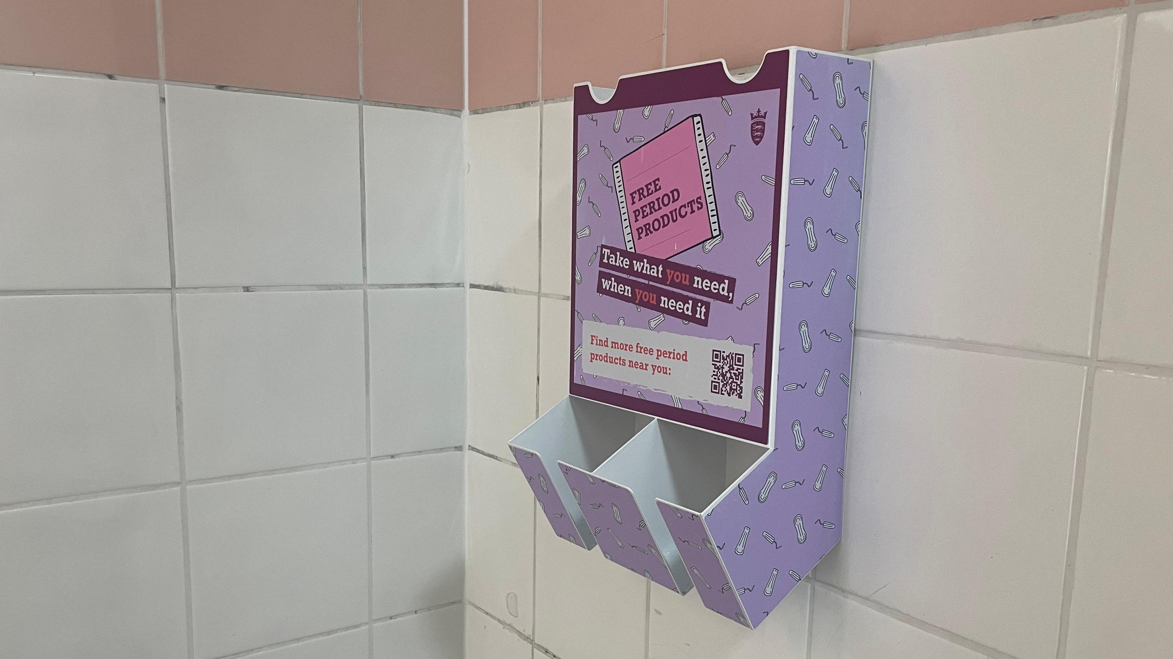 Wall-mounted dispensers for period products photographed in a Jersey public toilet. The dispenser is lilac in colour and the front reads: "Free period products." The dispenser has been mounted on a white tiled wall.