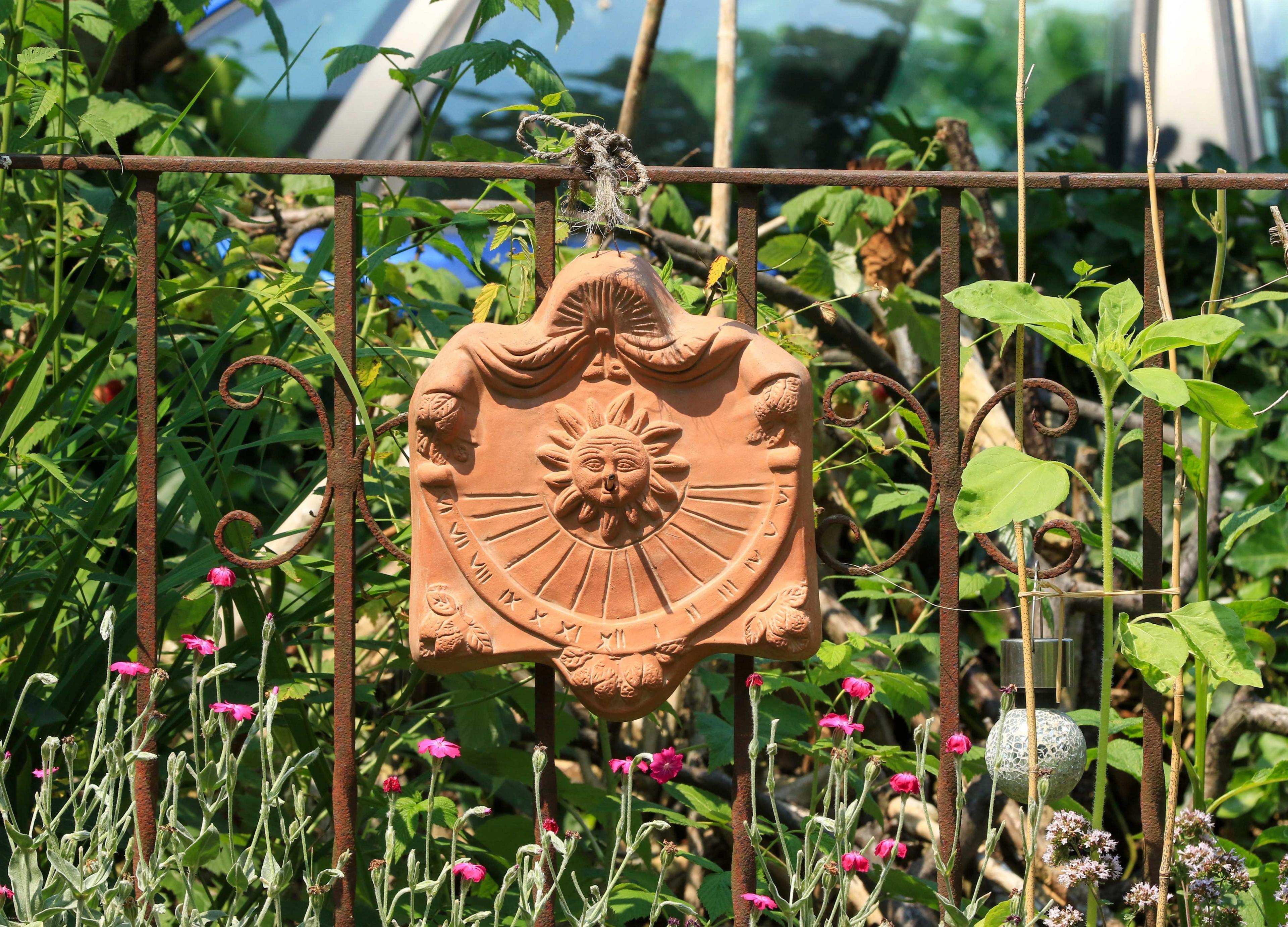 A clay sundial on metal railings. Wildflowers and greenery are behind the railings