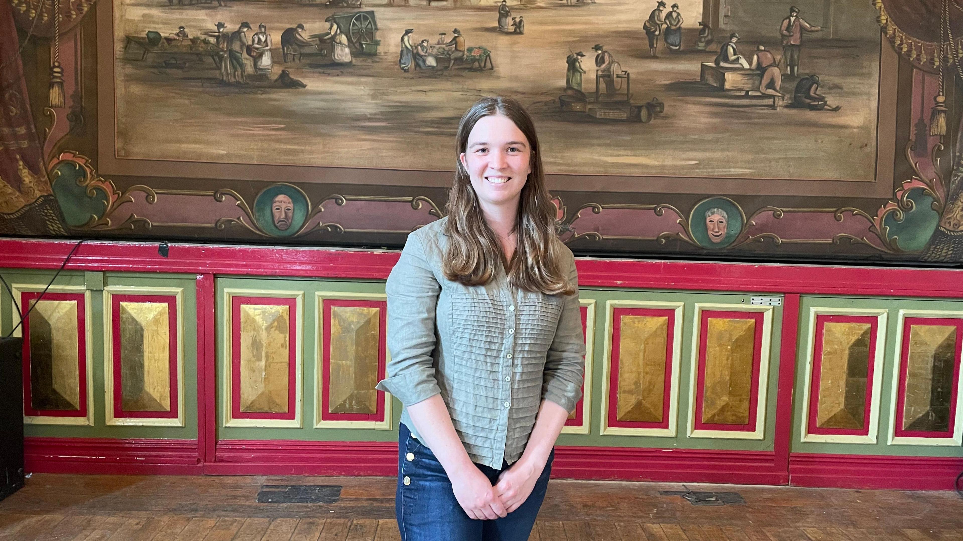 Josie Guthrie from Settle Music Hall standing in front of the stage at the hall.

Josie is smiling and has shoulder length brown hair and is wearing a light green shirt with blue jeans.

The theatre scenery depicts several people in the town of Settle at work.