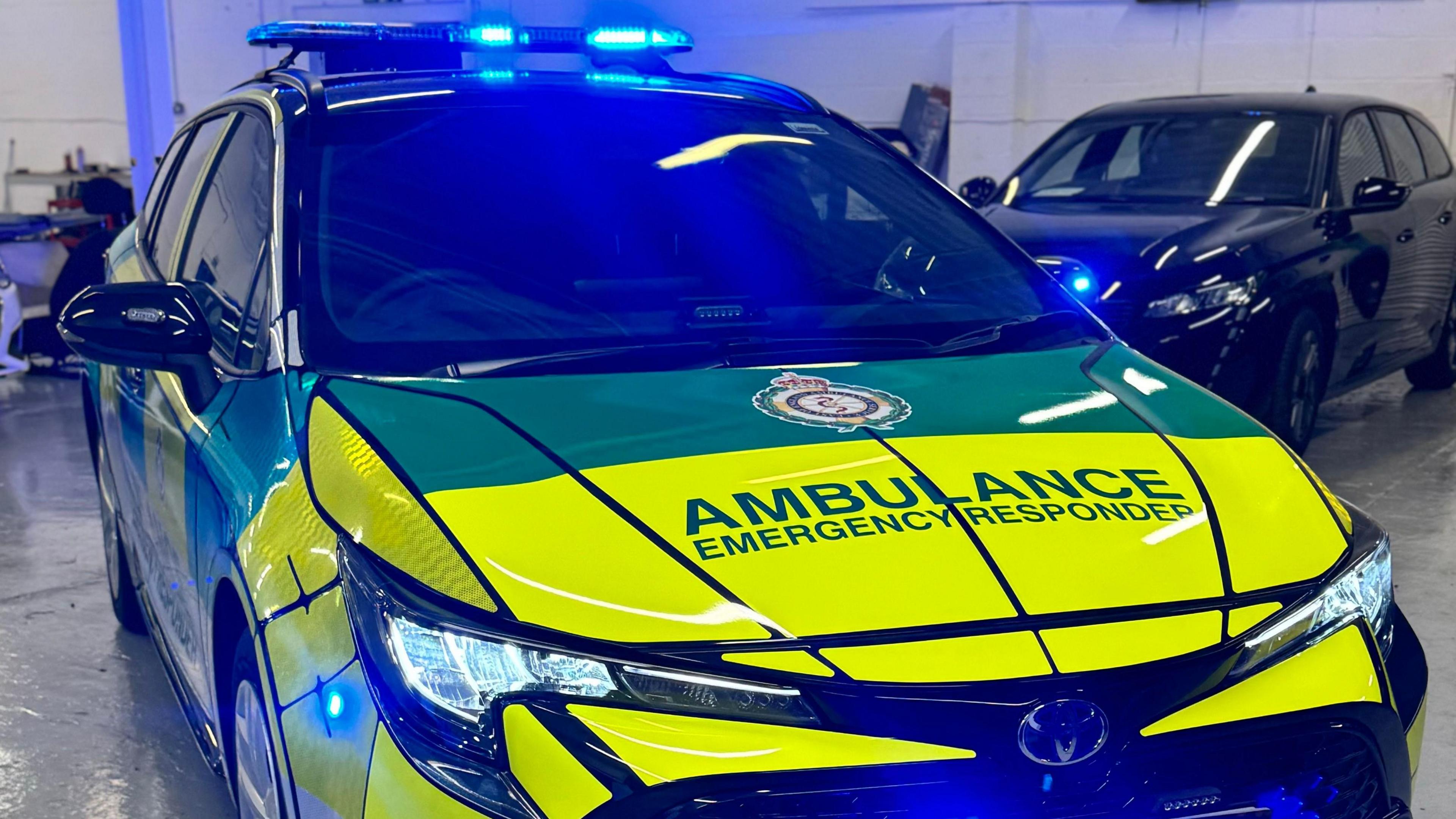 An ambulance, emergency response vehicle, in yellow and orange in a room, with two vehicles behind it. 