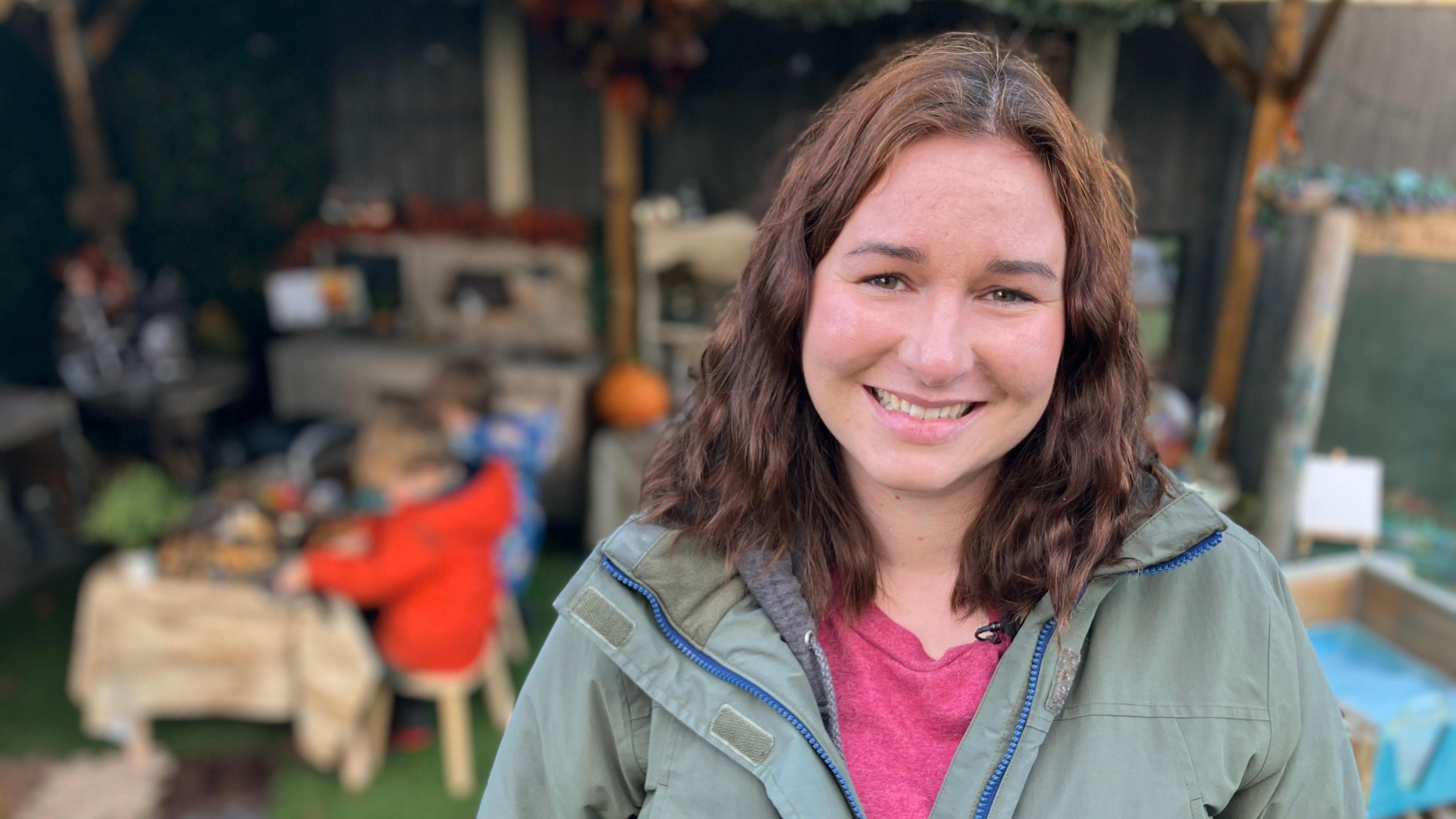 Georgina who has brown curly hair and is wearing a green coat and pink t-shirt is smiling at the camera. Behind her there are two children sitting a table outside busy playing.