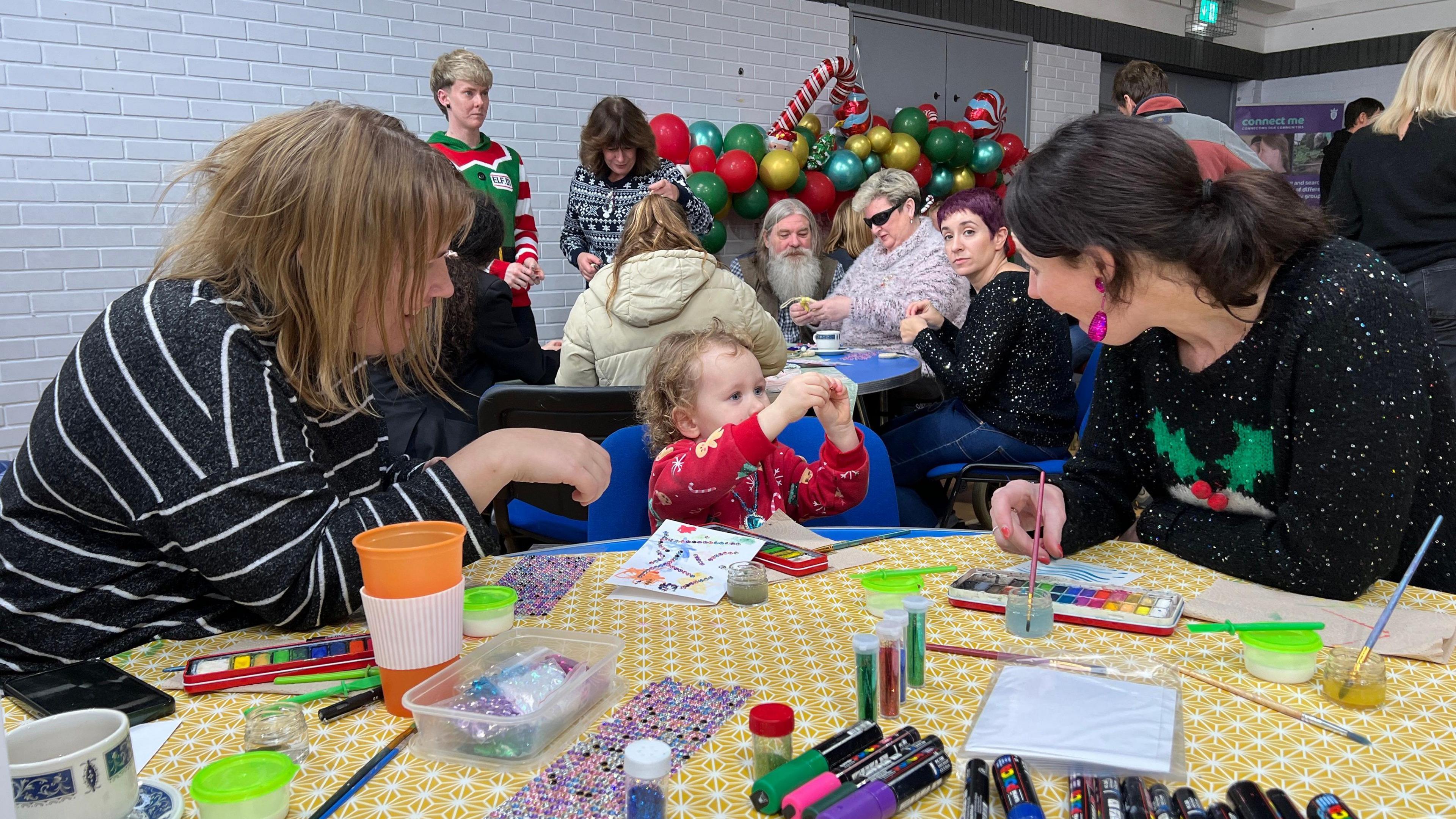 A little girl is sitting at a chair by a table between two women and she's showing one of them what they've made in their hands. The woman is reacting with surprise to the child. The child is wearing a red Christmas jumper with the woman to the right in a black Christmas jumper with a Christmas Pudding decoration. The other woman is wearing a dark jumper with white stripes. On their table are arts and crafts items.