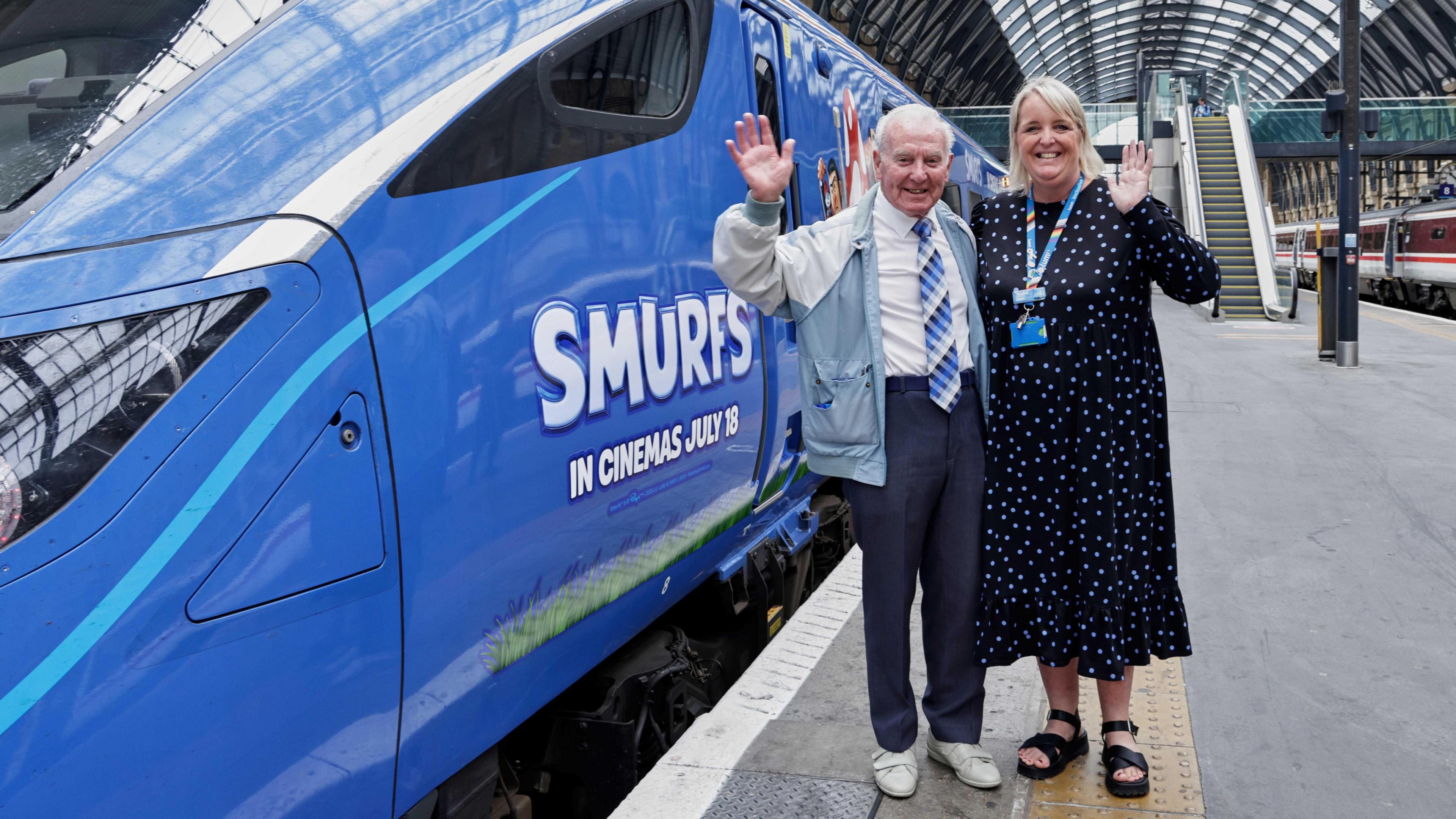 A man with short white hair, a white shirt, a blue patterned tie and dark trousers standing with a woman who has light shoulder-length hair, a blue patterned dress, a lanyard and sandals, next to a blue train with "Smurfs" branding on it. They are standing on a platform in a railway station and waving at the camera.