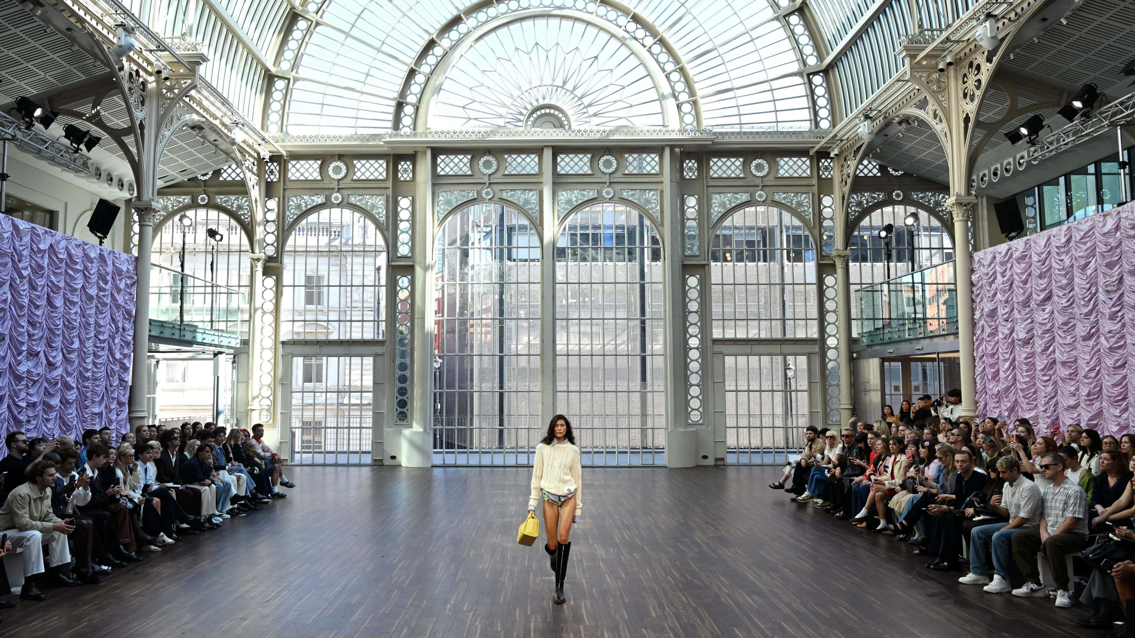A model walks a makeshift catwalk at in the Dorfman Conservatory at the Royal Opera House. The room is a large and grand glass conservatory.
