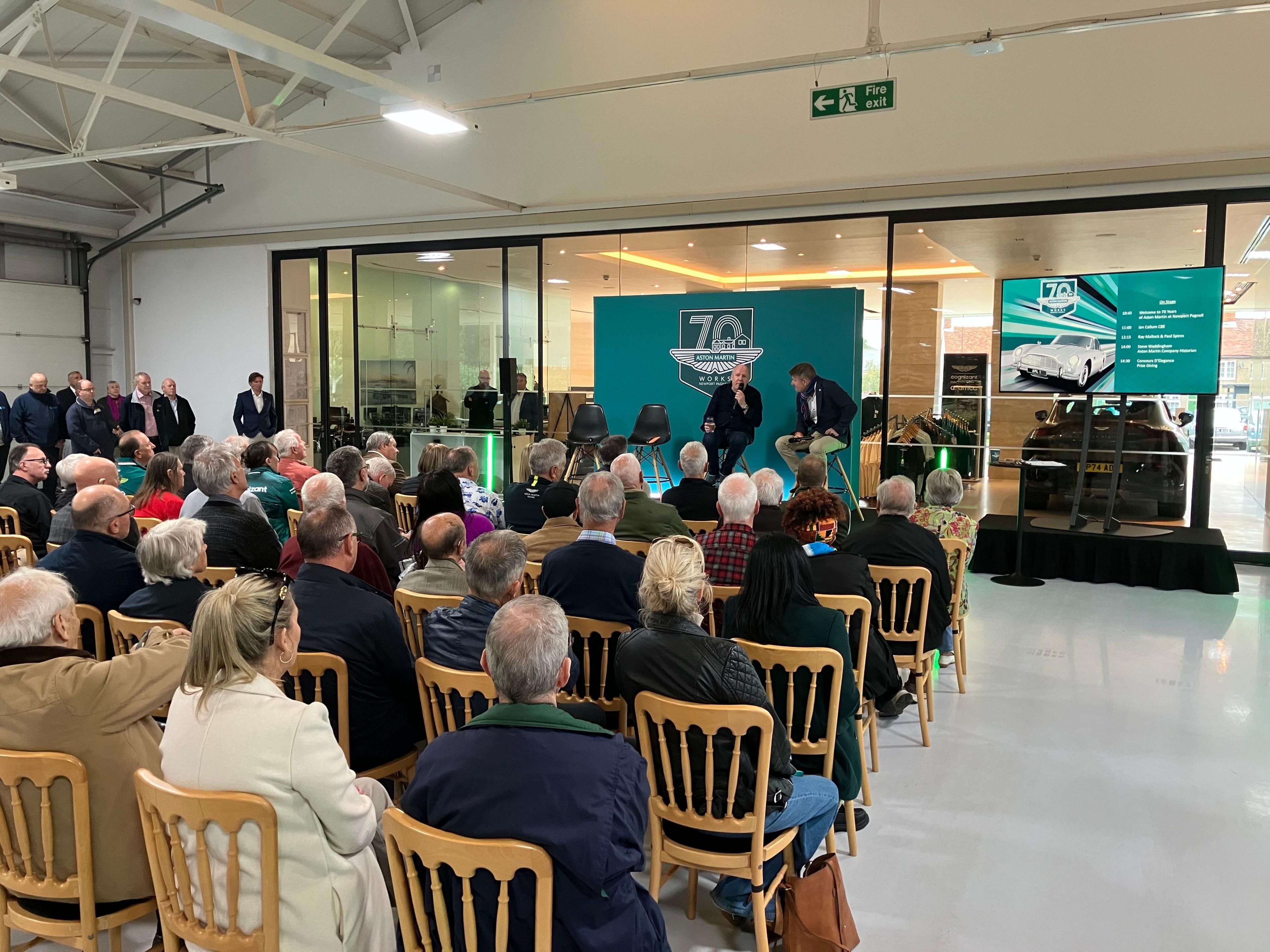 The photo is looking forward to a small stage where two people are sitting on stools with microphones. The number "70" can be seen with the Aston Martin logo behind them on a green background. The photo shows people sitting in six rows of chairs looking at the stage. 