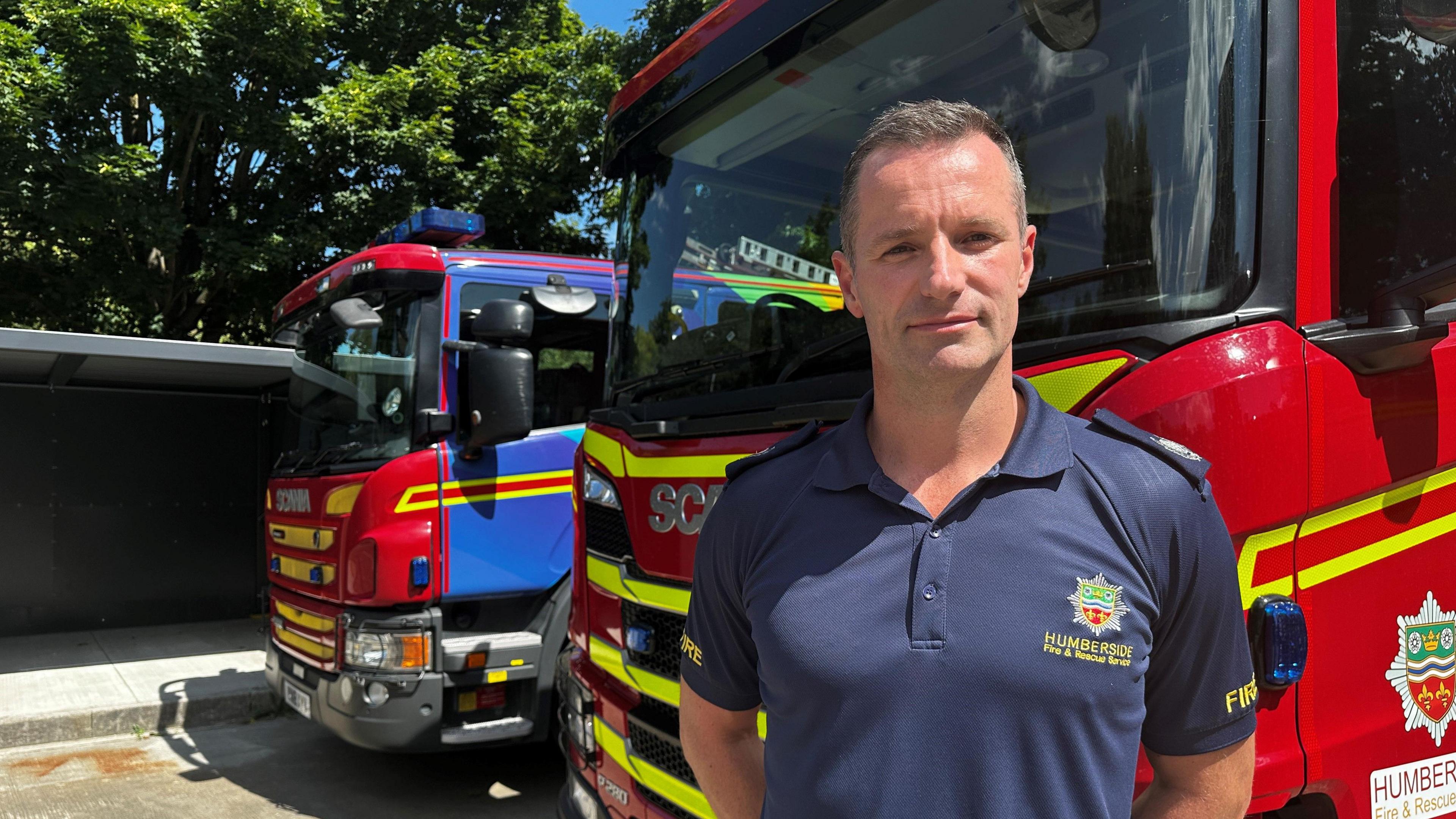 A man with short cropped hair stands in front of two red fire engines. He is wearing a dark blue shirt with a Humberside Fire and Rescue Service logo on it.