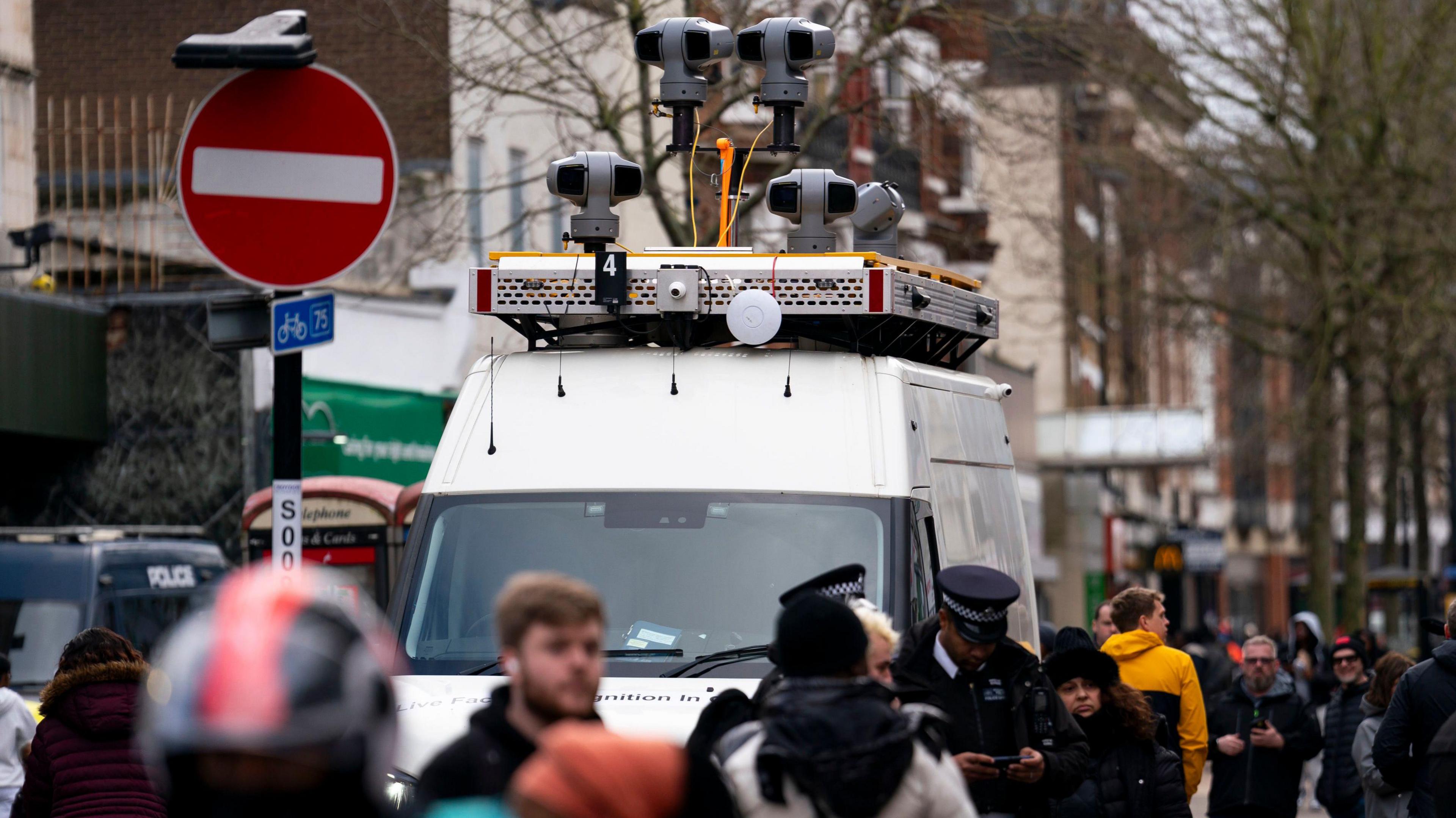 A live facial recognition van. It has about 5 visible multi-angle cameras on top of it. Some policemen stand around nearby and crowds of people from the public walk by too.