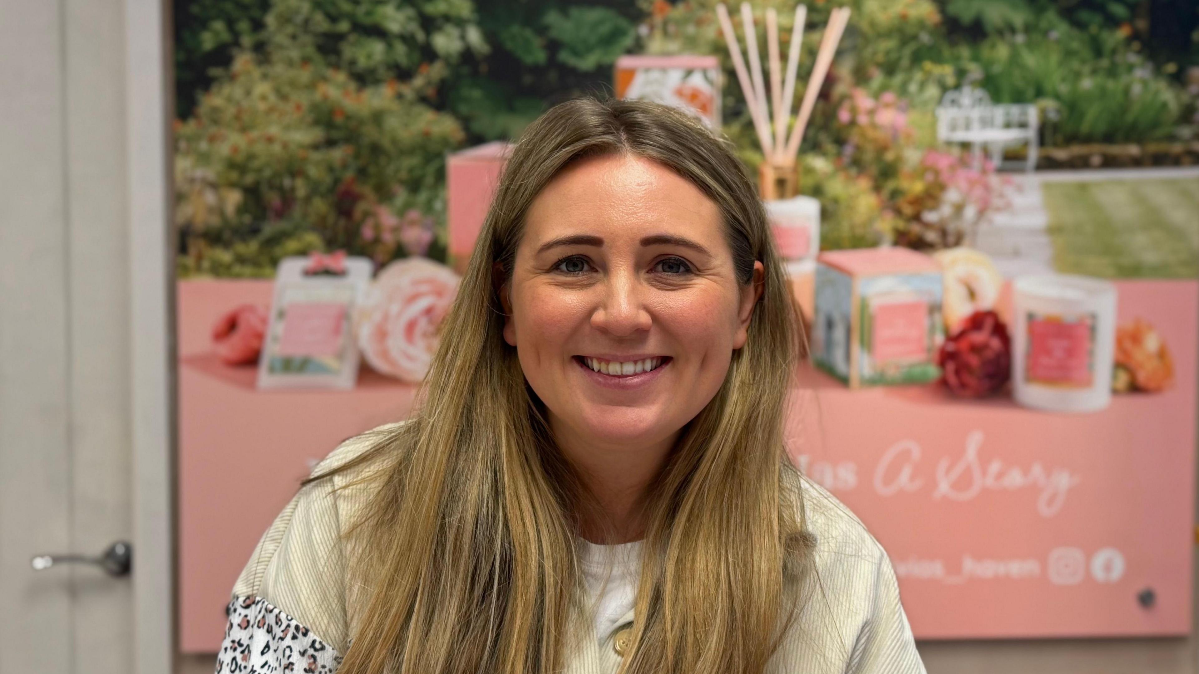 Woman wearing a tan shirt is smiling at the camera. She has blonde hair straight hair. There is a pink poster in the background with candles on it. 