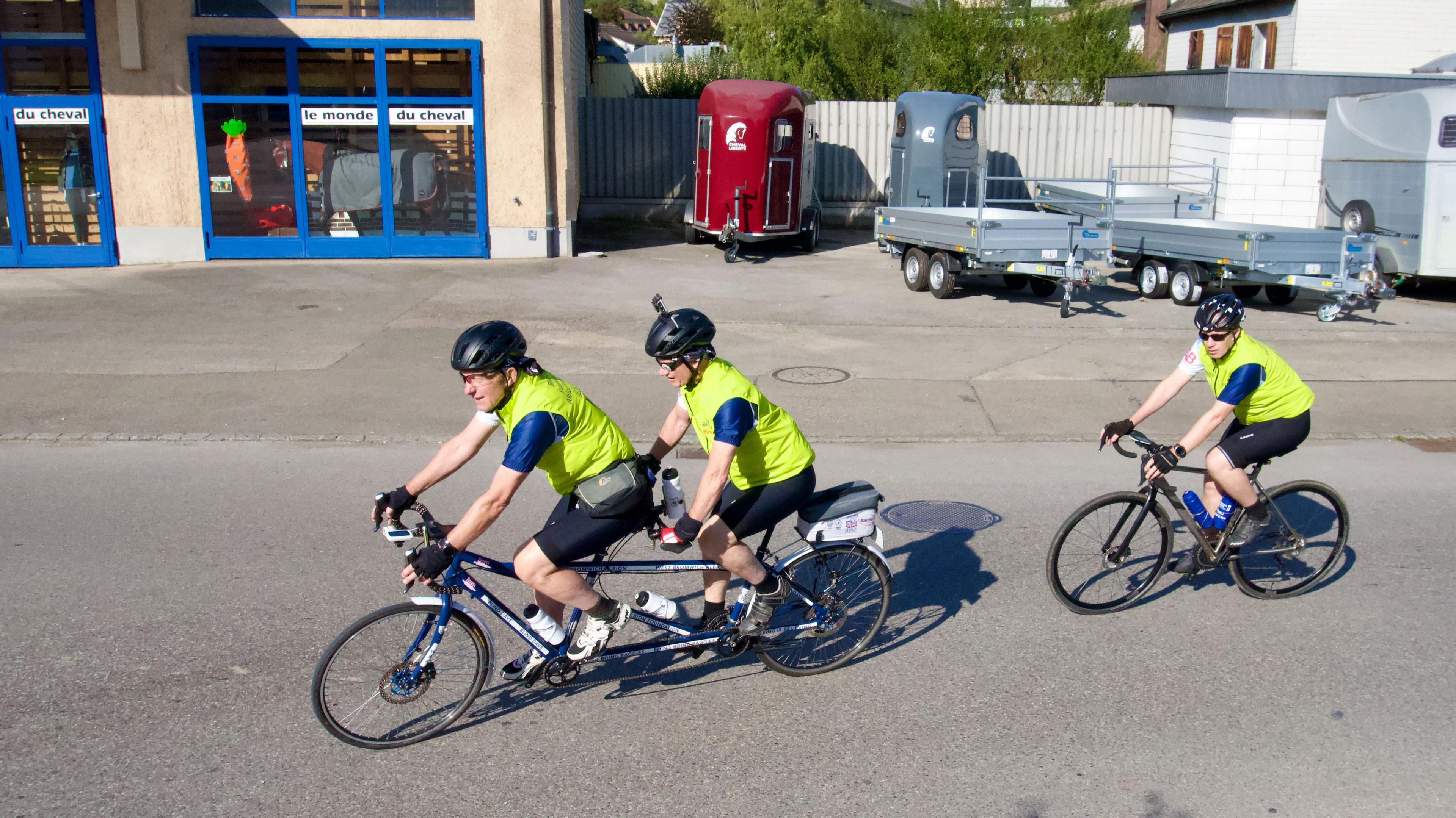 Three male cyclists, two on a tandem, cycle on a town centre road in the sunshine