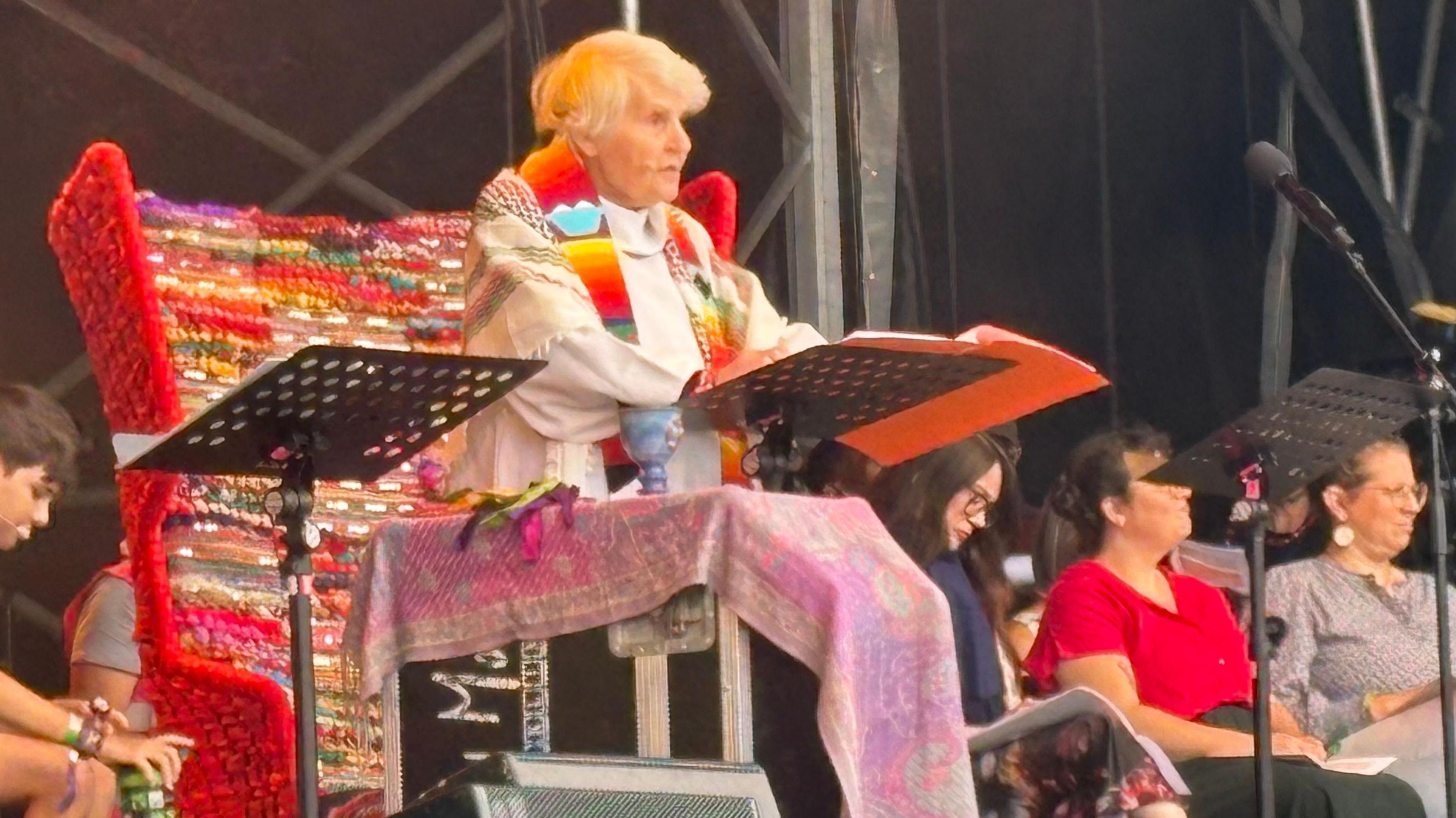 Sue Parfitt with medium-length white hair wearing a multi-coloured stole and white cloak. She is sitting on a red throne-like chair and reading from an orange booklet on a stand. There are people sitting to her left. The back of a festival stage is visible behind her.