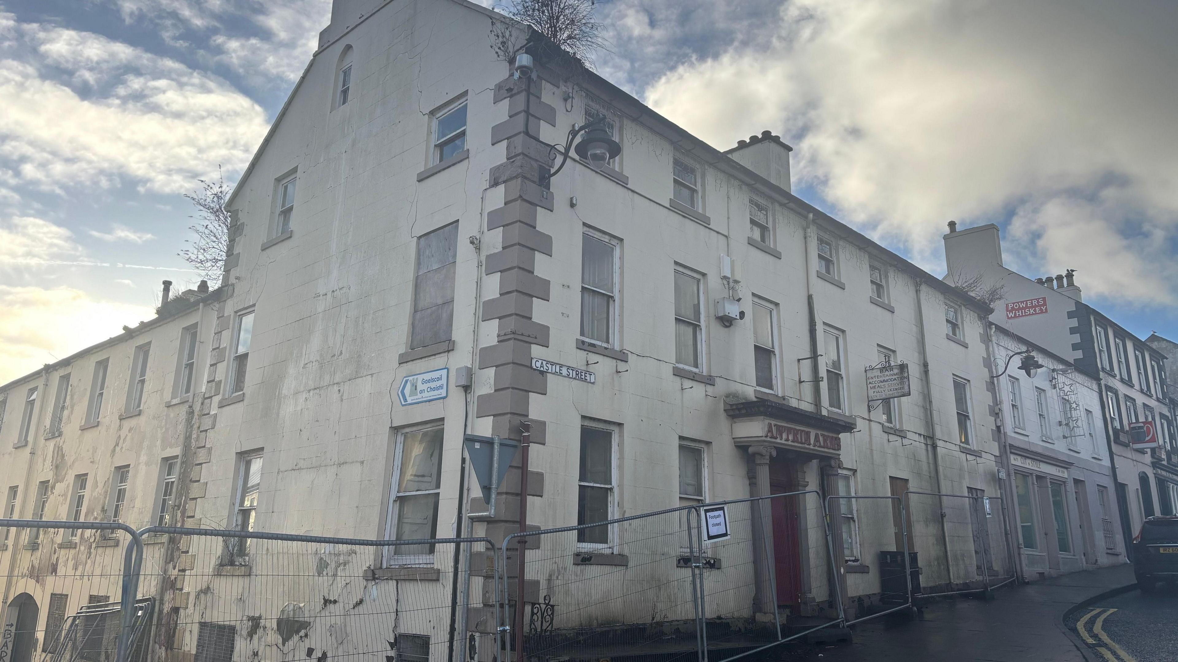 A side-on view of the former Antrim Arms hotel at the corner of Castle Street and Fairhill Street in Ballycastle in January 2025.  The building has a rendered exterior but the cream paint is fading and peeling in places.  Some vegation is growing out of the gutter on the roof and steel barriers are blocking off access to the building and adjoining street. 