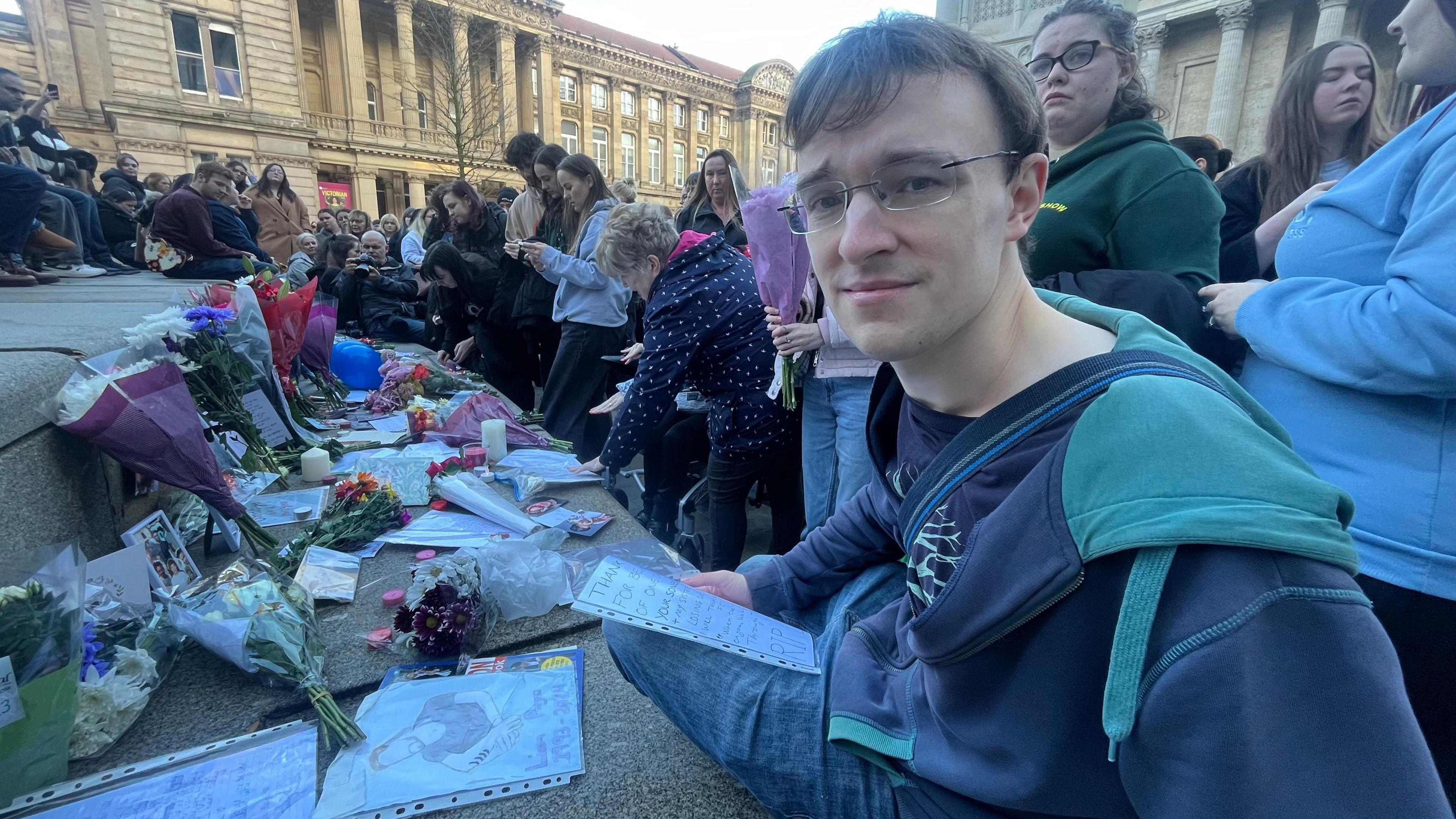 Jamie Parker stares at the camera while a crowd is behind him, and flowers and candles line a step next to him. He has brown hair and is wearing glasses and a navy blue zip-up jumper.