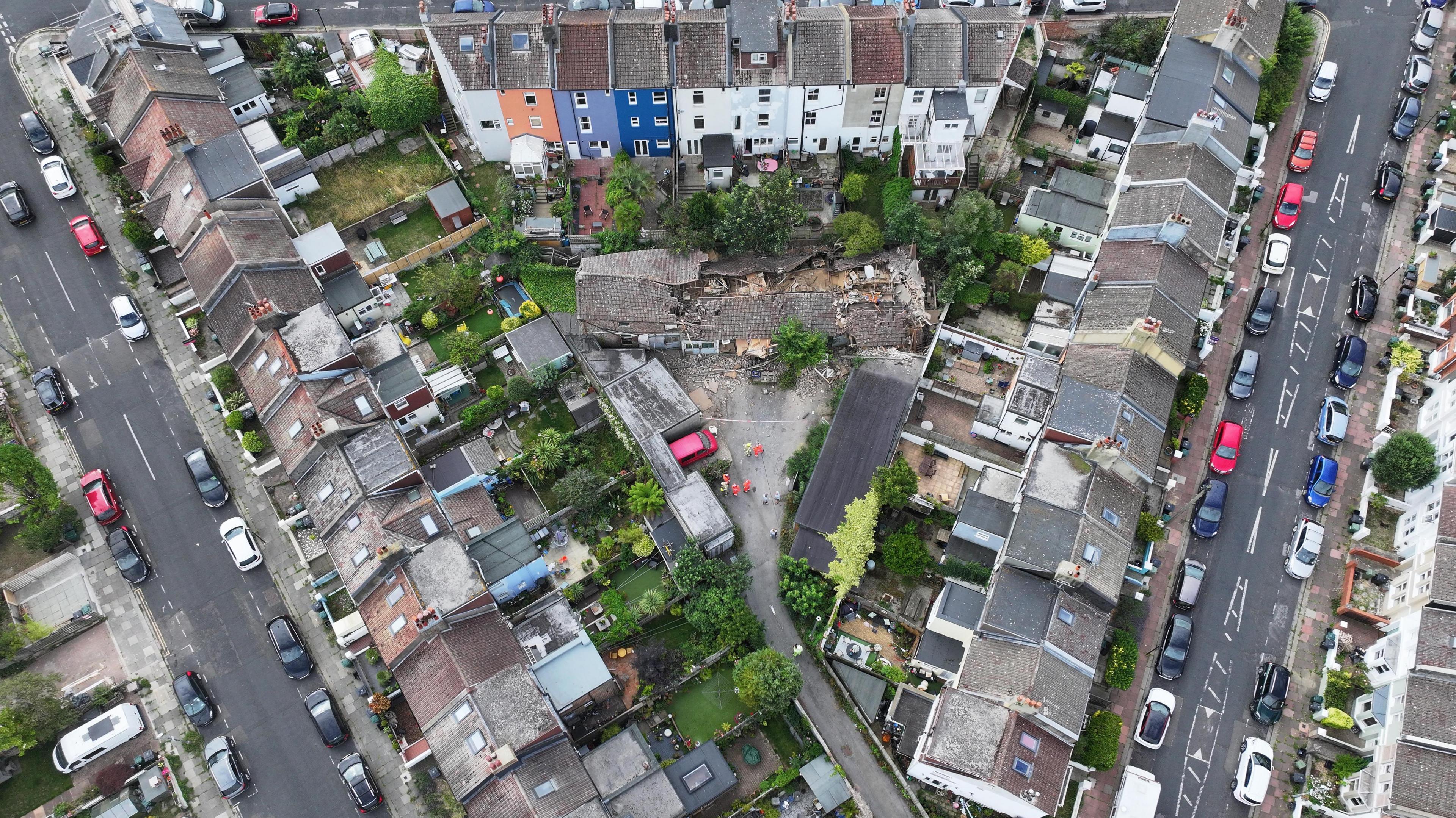 An aerial perspective of Ryde Road, Hartington Street and Carisbrooke Street in the Elm Grove area of Brighton. The houses that line the streets form a triangular formation around the site of the partially collapsed building. Back gardens face the affected site with fencing separating each property from the rest, as well as the site.
